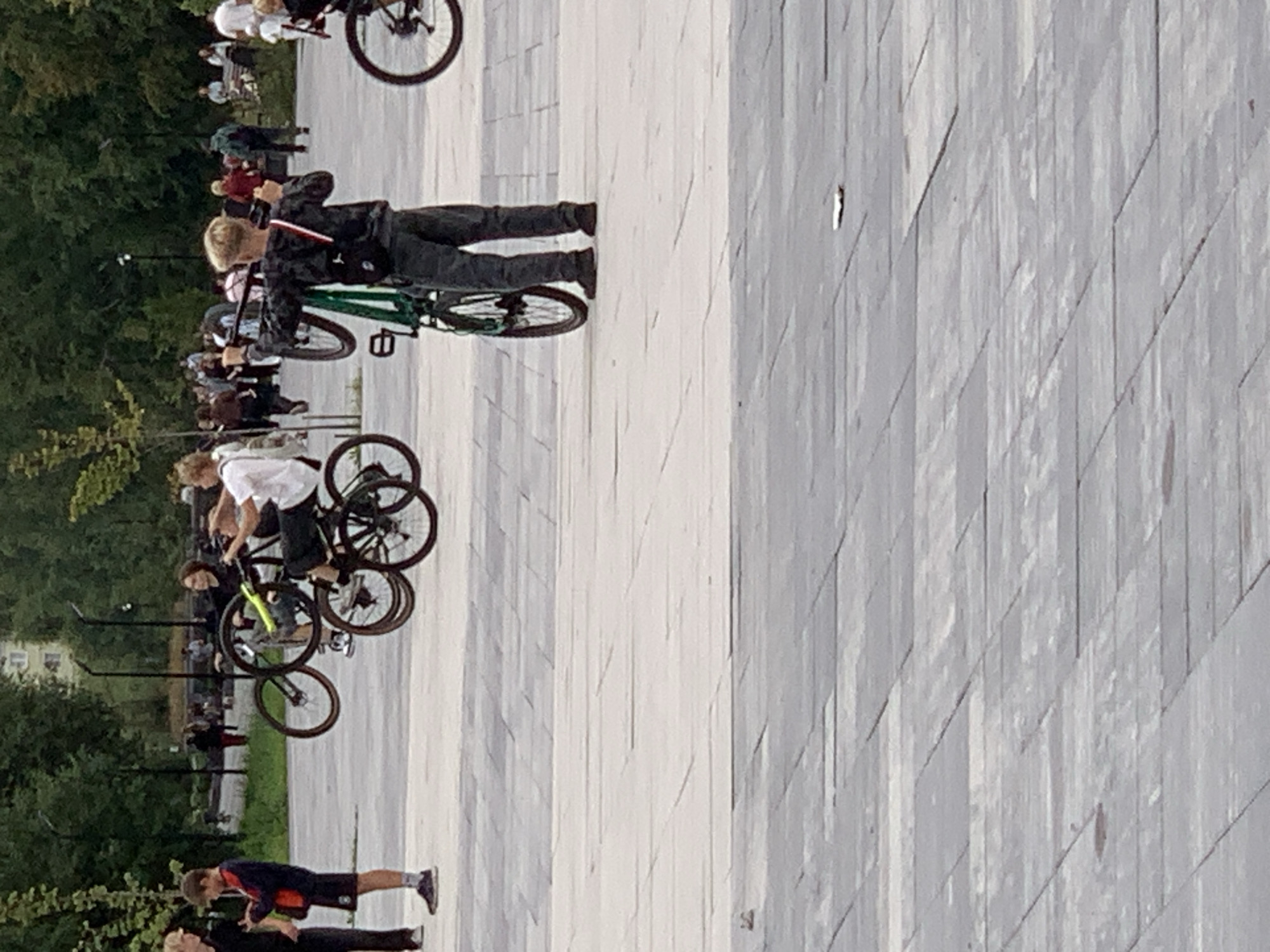 In an urban park setting with paved ground and scattered trees, several individuals are engaged with bicycles. In the foreground, a young person stands with a green bicycle, holding it upright with both hands. To the left, two other individuals are visible, one of whom is holding a bicycle. Further in the background, more people and bicycles are present, with some individuals appearing to interact with their bikes. The overall atmosphere suggests a casual gathering or activity involving bicycles. The location is stated to be Prokopyevsk, Russia.