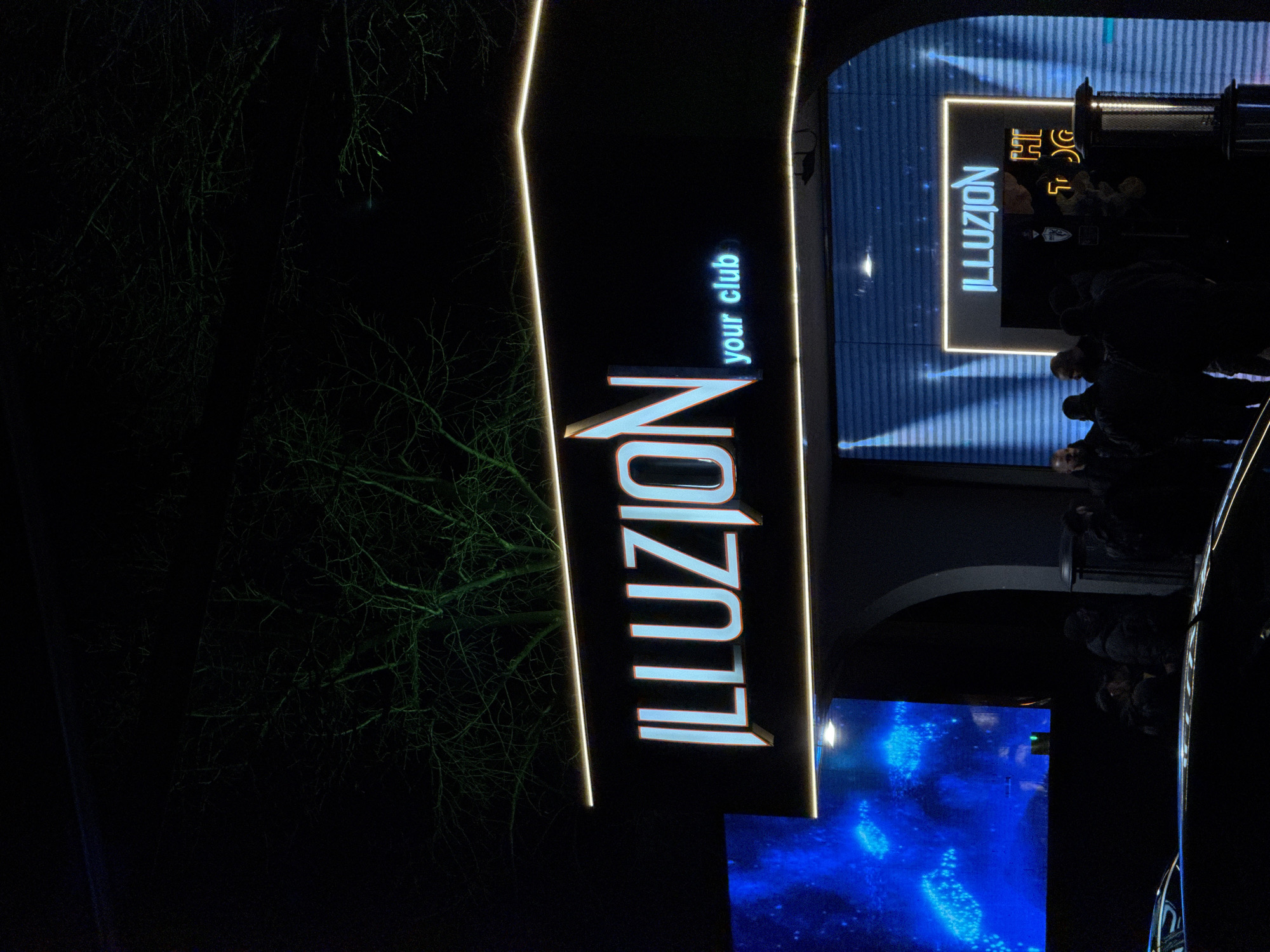 The image depicts the exterior of the "ILLUZION your club" nightclub in Sofia, Bulgaria. The club's illuminated sign is prominently displayed. Patrons are visible in silhouette near the entrance. A screen displays a blue graphic. Bare trees are visible in the background.