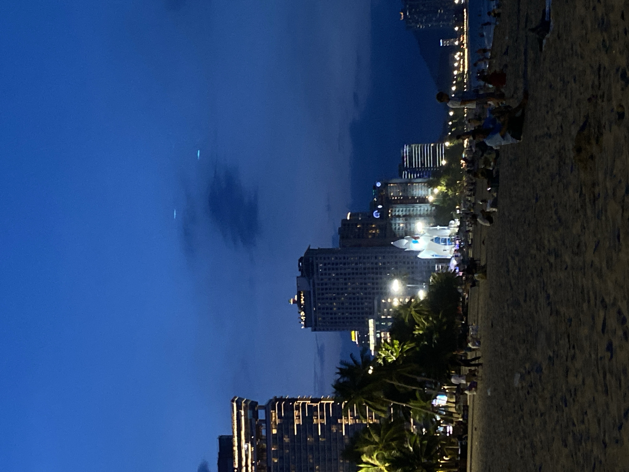 The image depicts a nighttime scene on a sandy beach in Nha Trang Ward, Vietnam. Tall buildings with illuminated windows line the coast, contrasting with the dark blue twilight sky. Palm trees add to the tropical atmosphere, their silhouettes visible against the lit structures. Several people are scattered across the beach, some walking, others sitting or standing, suggesting a relaxed evening gathering. The lighting from the buildings and streetlights creates a warm glow on the sand. The overall mood is serene and peaceful, with the gentle hum of the city providing a backdrop to the tranquil beach setting. The image captures the vibrant urban life of Nha Trang extending to its scenic coastline.