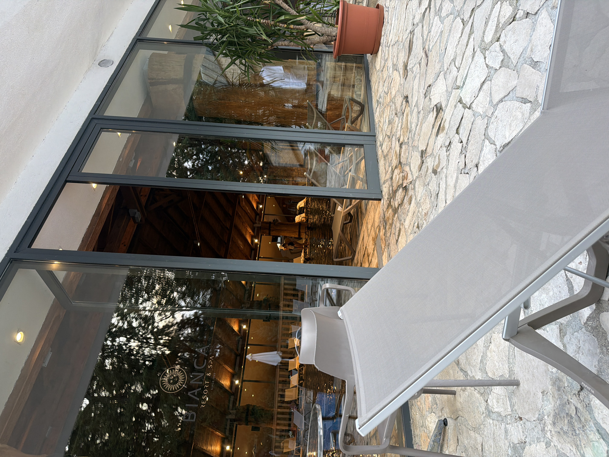 The image depicts the outdoor seating area of the "BIANCA RESORT & SPA SINCE 1949" in Kolašin, Montenegro. A grey lounge chair is in the foreground, angled towards the right. Behind it, a stone patio is visible, with various chairs and tables set up near a body of water, which appears to be a pool. Large glass doors and windows reflect trees and the interior of the resort, which features exposed wooden beams and warm lighting. A tall green plant in a terracotta pot stands to the right of the glass facade. Inside the resort, visible through the glass, are lounge chairs, a person in a white robe, and another person in swimwear. The overall impression is one of a tranquil, resort-like environment.