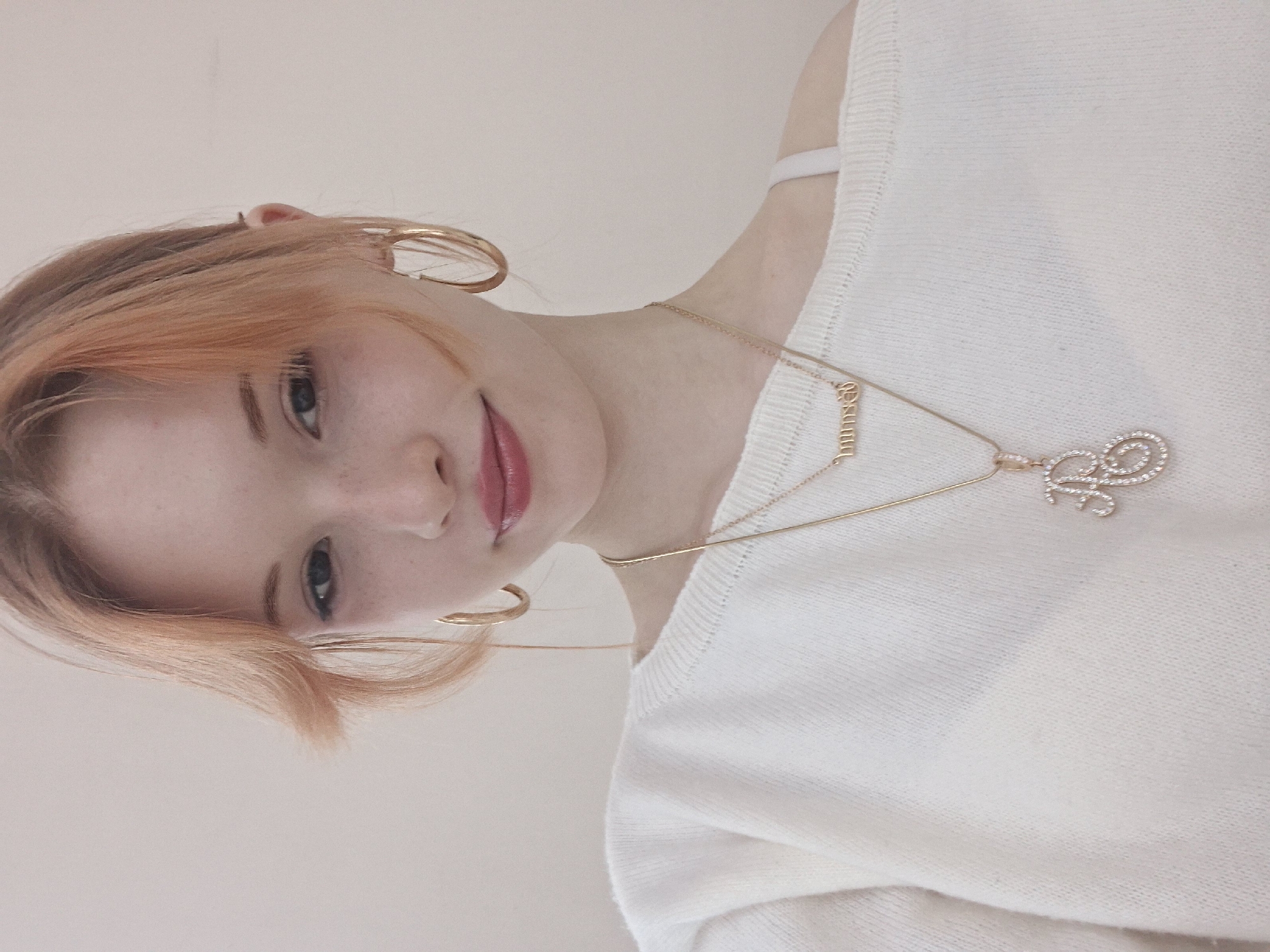 This media file features a young person, depicted from the chest up, against a plain, light-colored background. The image is rotated 90 degrees clockwise from what appears to be its original orientation, presenting the subject with their head towards the left side of the frame and body extending towards the bottom right.
The subject has light reddish-blonde hair, possibly dyed, with visible darker roots, and a fringe across their forehead. Their face is fair with scattered freckles, particularly visible around the nose. They have blue eyes and their lips are slightly parted, revealing a subtle smile enhanced by pink lip gloss.
They are wearing a white, lightly textured sweater. A white bra strap is visible on their right shoulder (which appears on the left side of the image as displayed). Several accessories are worn: large gold hoop earrings in both ears and two layered gold necklaces. One necklace features a small pendant spelling out "Jasmine" in block letters. The other, longer necklace, has a larger, ornate pendant encrusted with clear rhinestones, resembling a stylized letter, possibly an 'L' or a similar flowing script.
The activity captured is a direct gaze at the camera, suggesting a posed portrait. The setting is indoors, indicated by the plain wall and soft, even lighting, which makes the time of day indeterminate. No specific weather conditions or location cues are visible within the image to confirm the provided context of Chaikovsky, Russia. The visible text is the name "Jasmine" on one of the necklaces and the stylized initial on the other. The overall mood is serene and personal.