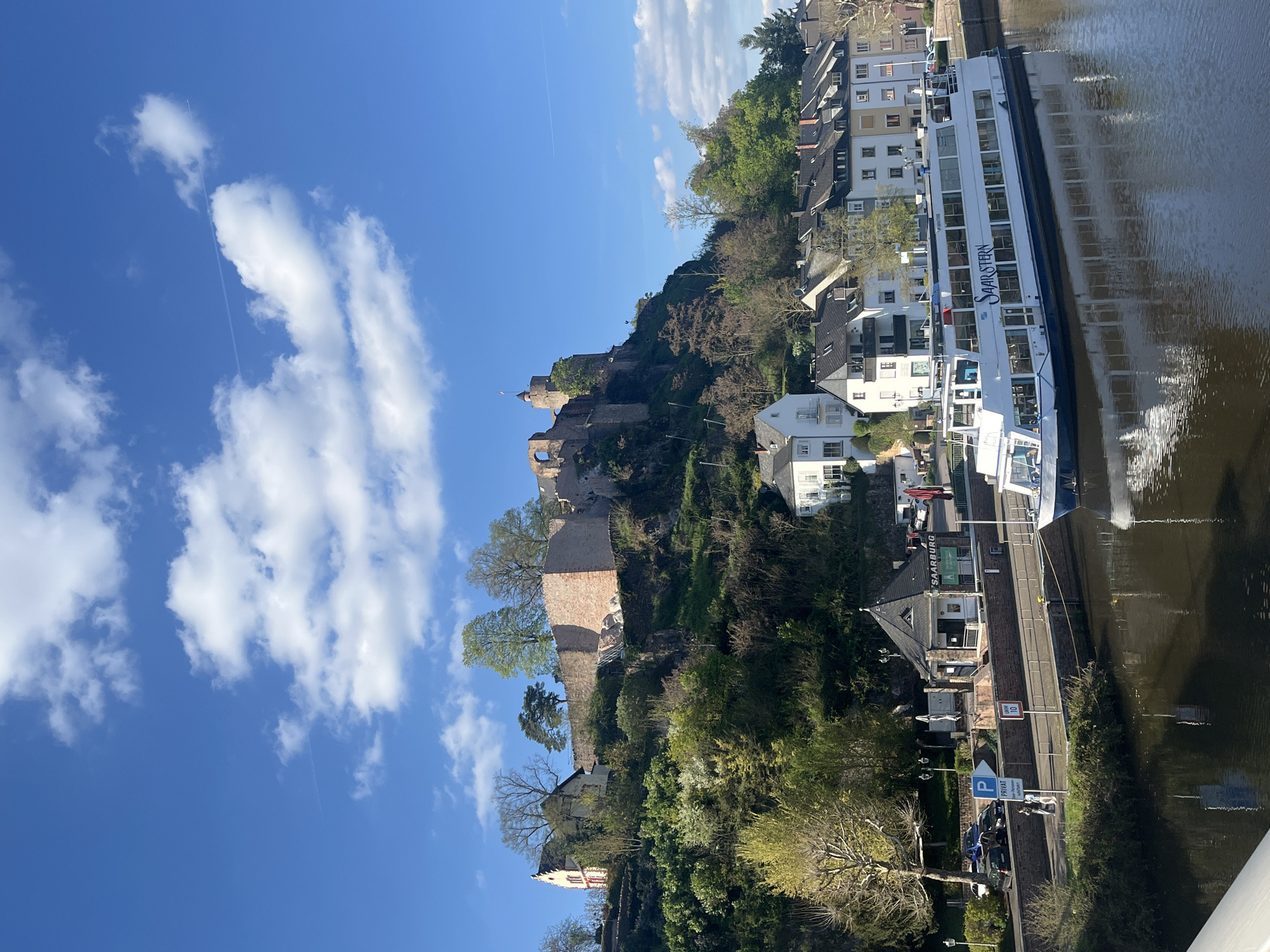 The image depicts a sunny day in Saarburg, Germany, with a view of the town and the Saar River. A large river cruise ship, the "Saarstern," is docked along the riverbank. Above the river, the town is built on a hillside, topped by Saarburg Castle. The castle is partially in ruins, with some sections of the walls and towers still standing. The hillside is covered with trees and vegetation, interspersed with houses and buildings of various architectural styles. A road runs along the riverbank next to the docked ship, with cars and a parking sign visible. The sky is blue with scattered white clouds and an airplane trail.