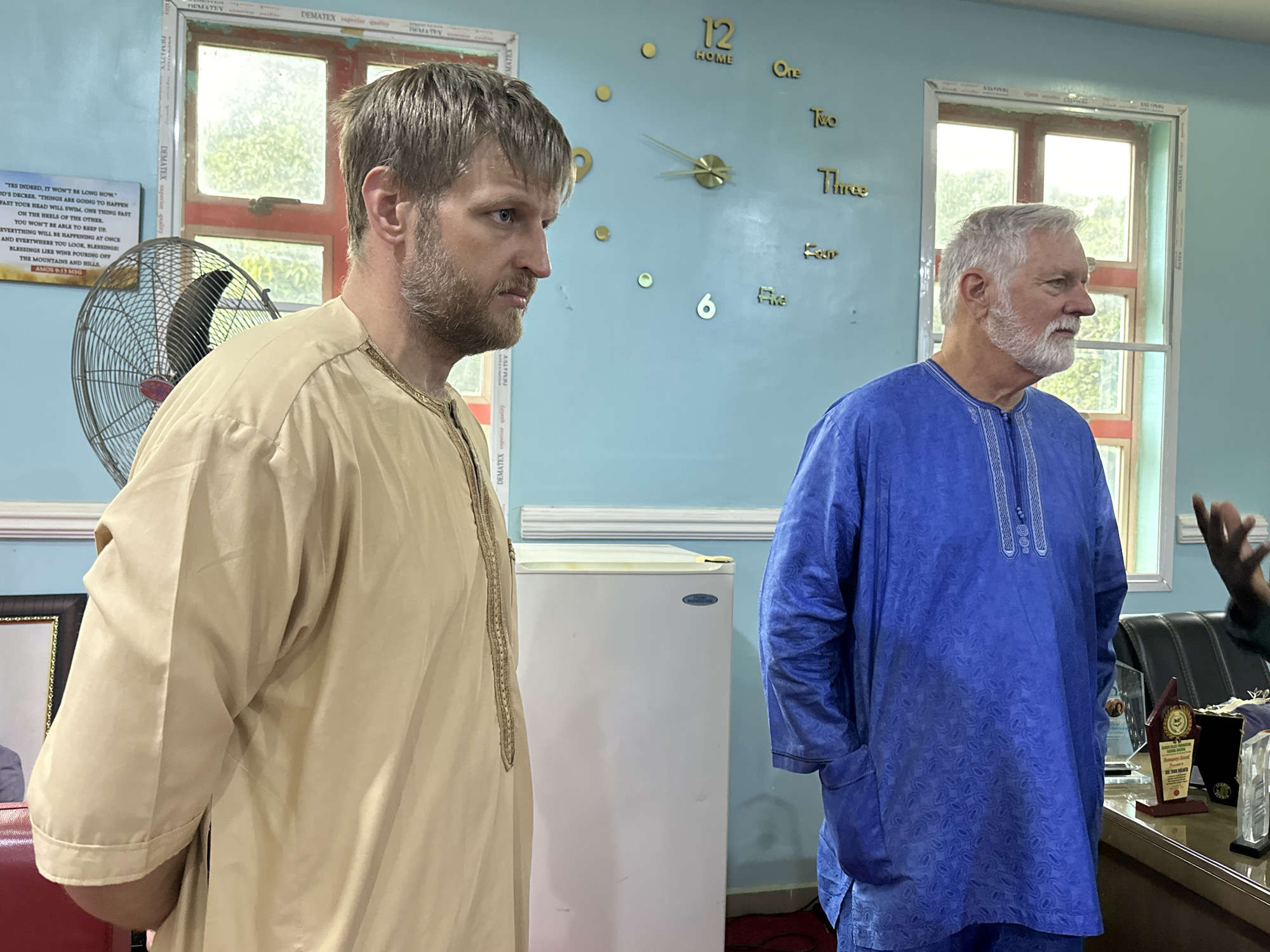 In Kakau, Nigeria, two men stand in an indoor room with light blue walls and two windows that reveal trees outside. A large, gold, 3D clock is mounted on the wall, with the numbers 12, 1, 2, 3, 4, 5, and 6 visible. To the left, a black electric fan is partially obscured by a man wearing a tan robe with gold trim. He has blonde hair and a beard, and he is looking to his right. In front of him is a framed picture. To his right, another man with white hair and a white beard wears a blue robe with a patterned design. He is looking to his right, with his hands in his pockets. Between the two men is a small, white refrigerator. On a desk behind the man in the blue robe, several awards are displayed, including a wooden plaque and a glass trophy.