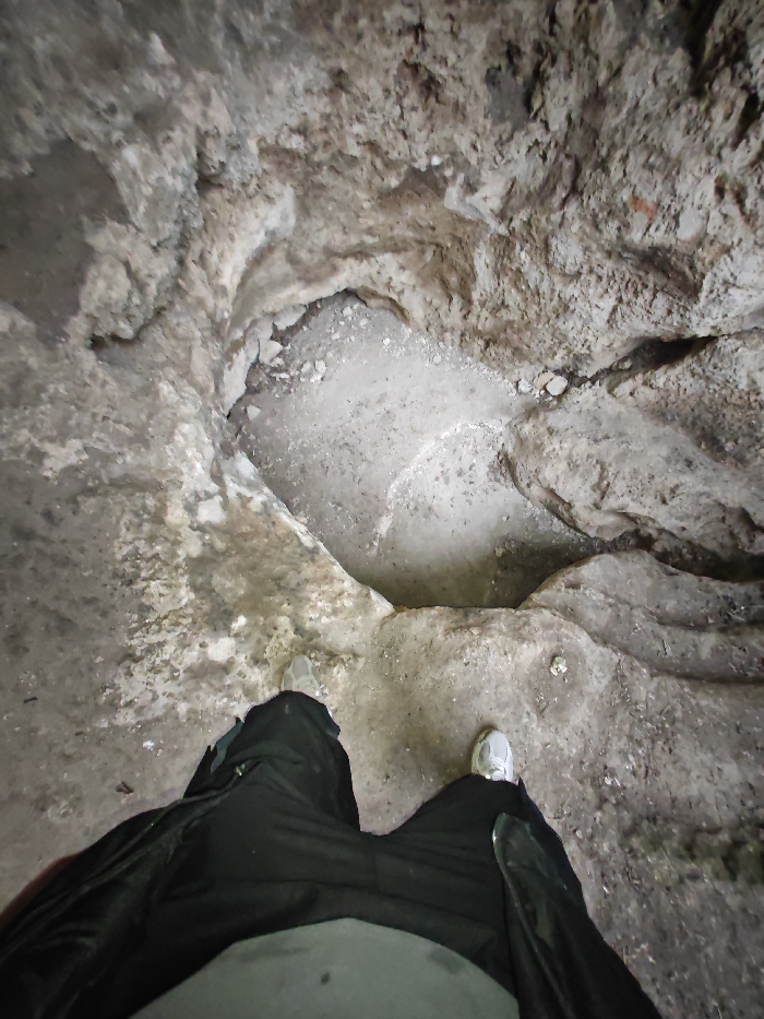 The image depicts a first-person, high-angle view looking down into a subterranean or rock-cut environment, consistent with a natural cave or ancient excavation site within the Shumen Plateau region near Shumen, Bulgaria. The foreground is dominated by the lower torso and legs of an individual wearing dark, possibly black, trousers and light grey or white athletic shoes with white soles. The individual's feet are positioned on uneven, light grey and brown textured rocky ground. Directly ahead and below the viewer's position, a natural or excavated depression, approximately oval or heart-shaped, is visible. This depression, measuring roughly 1.5 to 2 meters in its widest dimension, has irregular, worn edges composed of the same textured rock. The bottom of the depression is filled with lighter, fine-grained material, appearing as compacted dirt, sand, or crushed rock, speckled with larger debris. The surrounding rock walls are uneven, coarse, and display varying shades of grey, brown, and off-white, suggesting erosion, mineral deposits, or exposed bedrock. No other individuals or distinct man-made objects are present. The scene is illuminated by ambient light, indicative of an enclosed or partially shaded space.