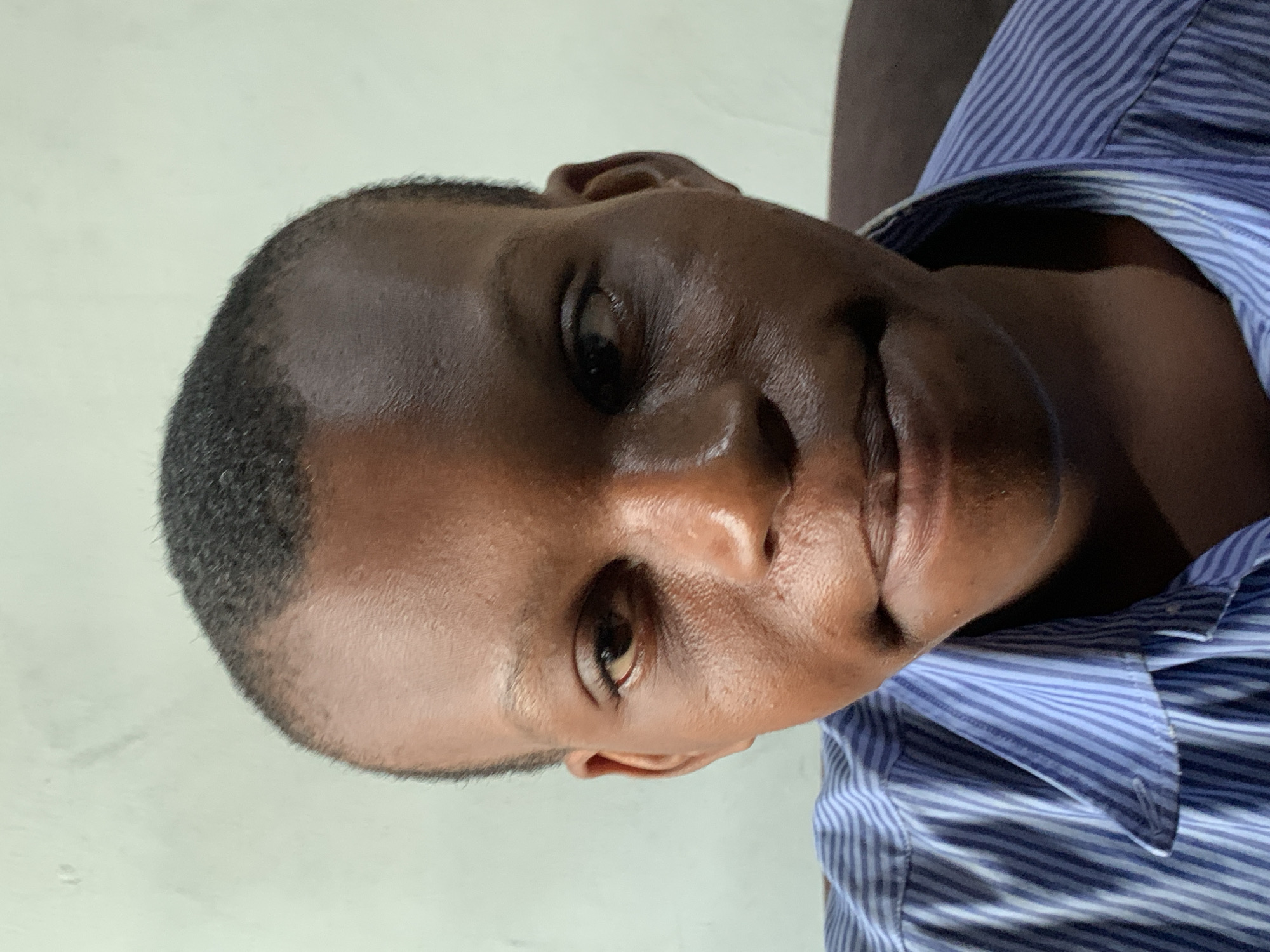 A close-up, portrait-style image, rotated 90 degrees clockwise, features a person, likely a man, with dark skin and short, closely cropped dark hair. The individual is looking directly towards the viewer with a neutral to slightly contemplative expression. Their eyes are dark and open.
The person is wearing a blue and white vertically striped collared shirt, visible on the right side of the frame (when viewed in its rotated state). The background is a plain, light-colored surface, possibly a wall, providing a simple backdrop. The lighting appears even, illuminating the subject's face, which shows some natural sheen on the forehead and nose.
No specific activity is taking place other than the person being photographed. There are no visible objects other than the individual and their clothing. No text is present in the image. The location context is Ibasa, Nigeria. The time of day cannot be definitively determined from the lighting, but it appears to be indirect or indoor illumination.