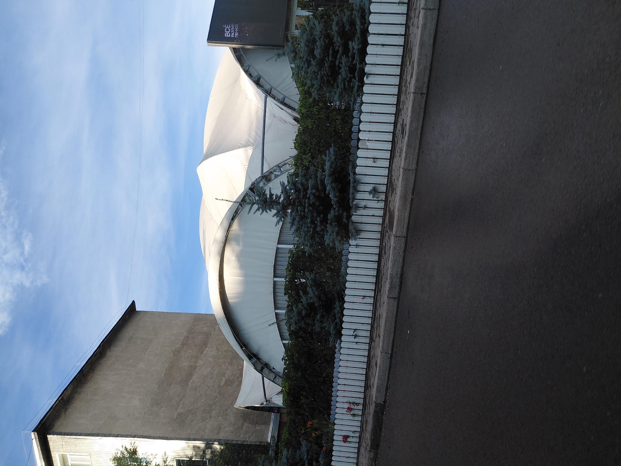 The image depicts a modern white tent-like structure with arched supports, likely a sports or event hall, located in the city of Karaganda, Kazakhstan. Adjacent to the tent is a tall, grey, multi-story building with visible windows. In the foreground, a low, white picket fence runs along a sidewalk. Several coniferous trees, appearing to be blue spruce, are planted behind the fence, with some visible flowering plants. The road in front of the sidewalk is wet, suggesting recent rain. The sky above is blue with scattered white clouds. A dark sign with white text is visible on the right side of the tent structure.