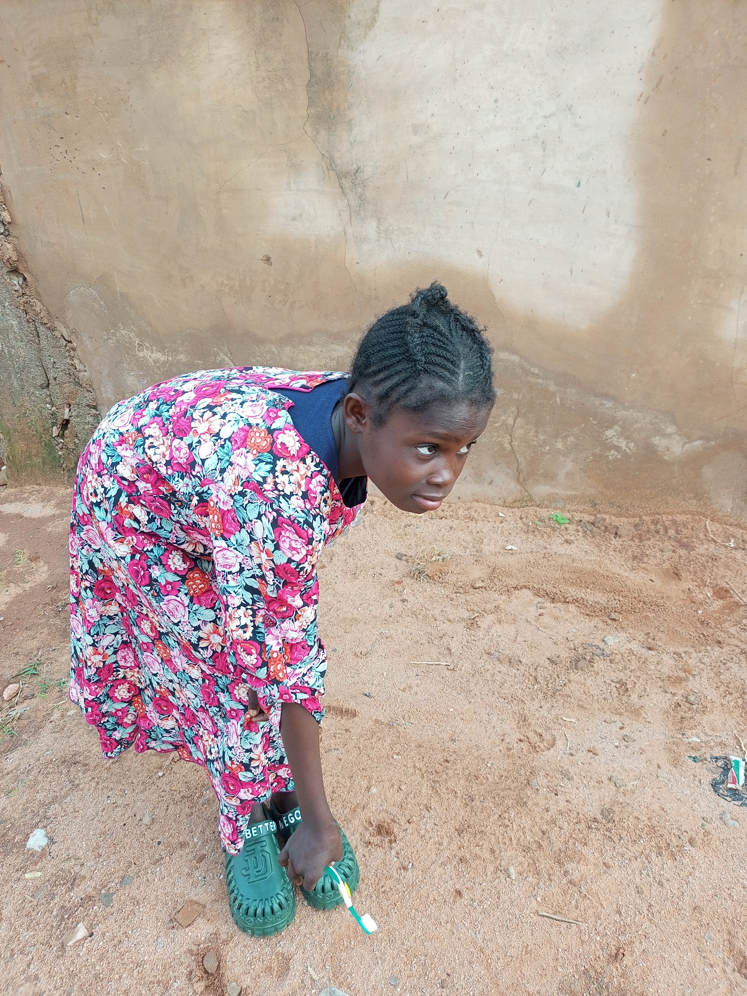 A young Black girl with braided hair and a floral dress bends over in a sandy, outdoor area with a textured, light-brown wall behind her. She is wearing green, open-toed shoes with the words "BETTER GO" visible on one of them. In her right hand, she holds a green toothbrush with white and yellow bristles. The ground is composed of sand and small pebbles, with a few dry twigs scattered about. The wall behind her appears weathered and cracked, suggesting an aged structure. The lighting is natural, casting shadows that indicate an outdoor environment in Tempure, Nigeria.
