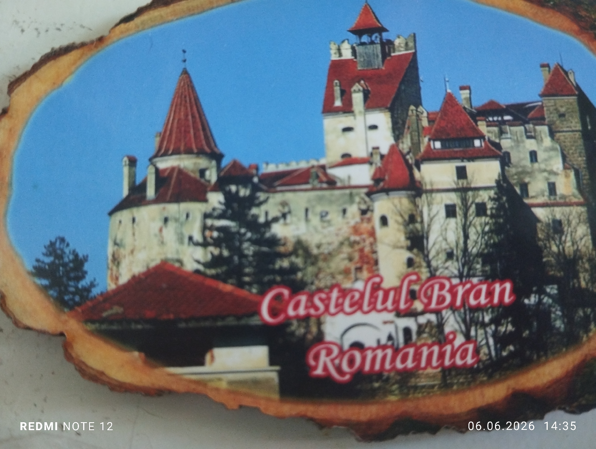 The image displays a wooden plaque with an irregular, natural bark-like edge, depicting a colorful image of a castle under a clear blue sky. The castle, identified by the superimposed red and white text as "Castelul Bran Romania," features multiple towers with distinct red conical and pitched roofs, light-colored stone walls, and numerous windows. Dark trees are visible around and partially obscuring parts of the castle structure. The wooden plaque rests on a plain, light-colored surface. A digital watermark "REDMI NOTE 12" is present in the bottom left corner of the overall photograph, and a timestamp "06.06.2026 14:35" is in the bottom right. There is no information within the image to indicate a location in the city of Burgas, Bulgaria; the depicted subject is Castelul Bran in Romania.