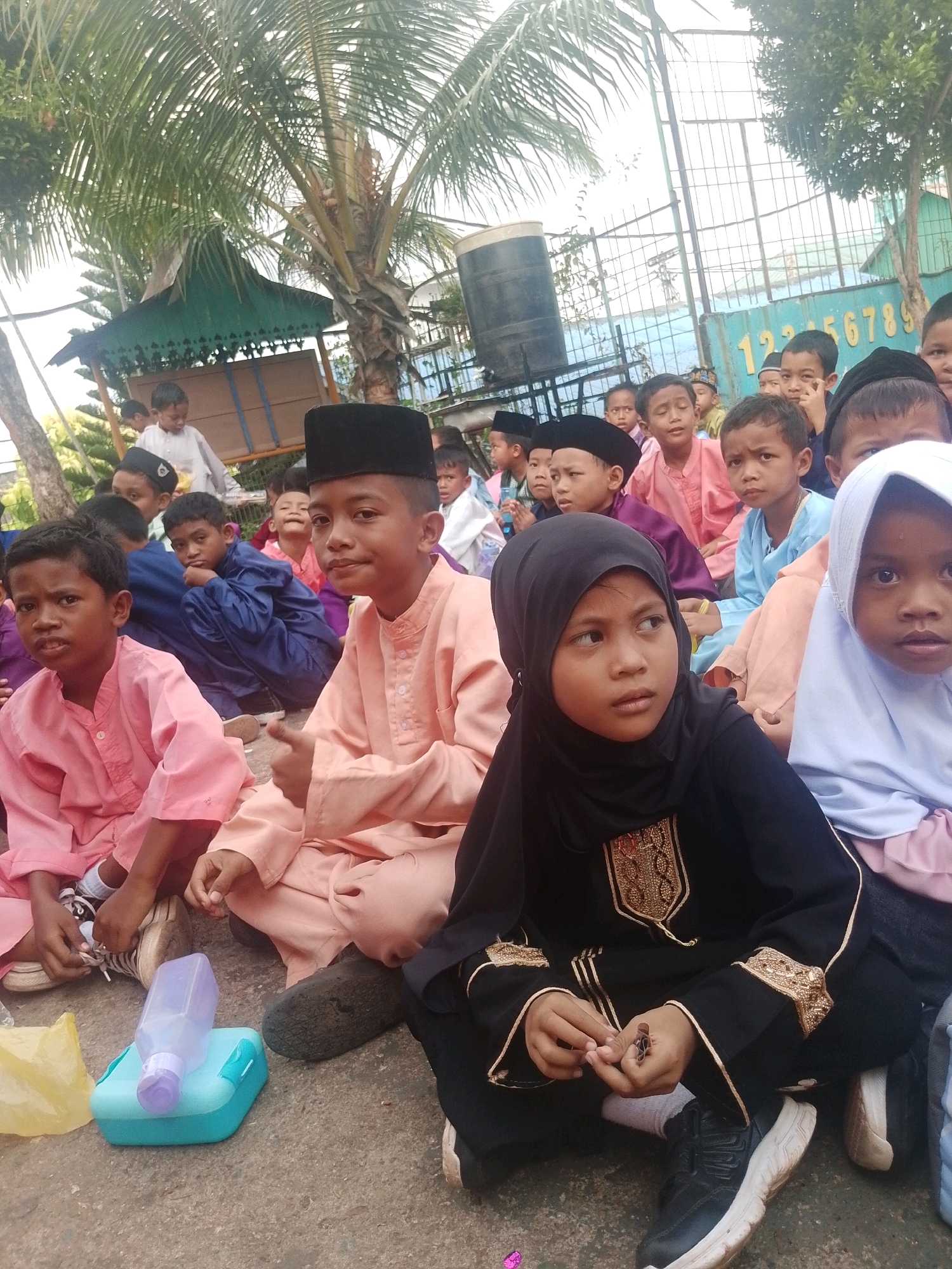 The image depicts a group of children, primarily young boys and a few girls, sitting on the ground outdoors. They appear to be dressed in traditional attire, with many boys wearing peci hats. Some children are looking towards the camera, while others are engaged in their own activities. In the foreground, a boy in a light pink outfit is tying his shoe, and a blue and purple lunchbox with a water bottle sits nearby. The background reveals a fence with the numbers "123456789" visible on a blue panel. Palm trees and a green awning suggest a tropical or sub-tropical outdoor setting, potentially a schoolyard or community gathering area. The presence of the fence and the numbers suggests a location like a school or sports facility. Based on the visual cues and context, this scene is likely located in Batam, Indonesia.