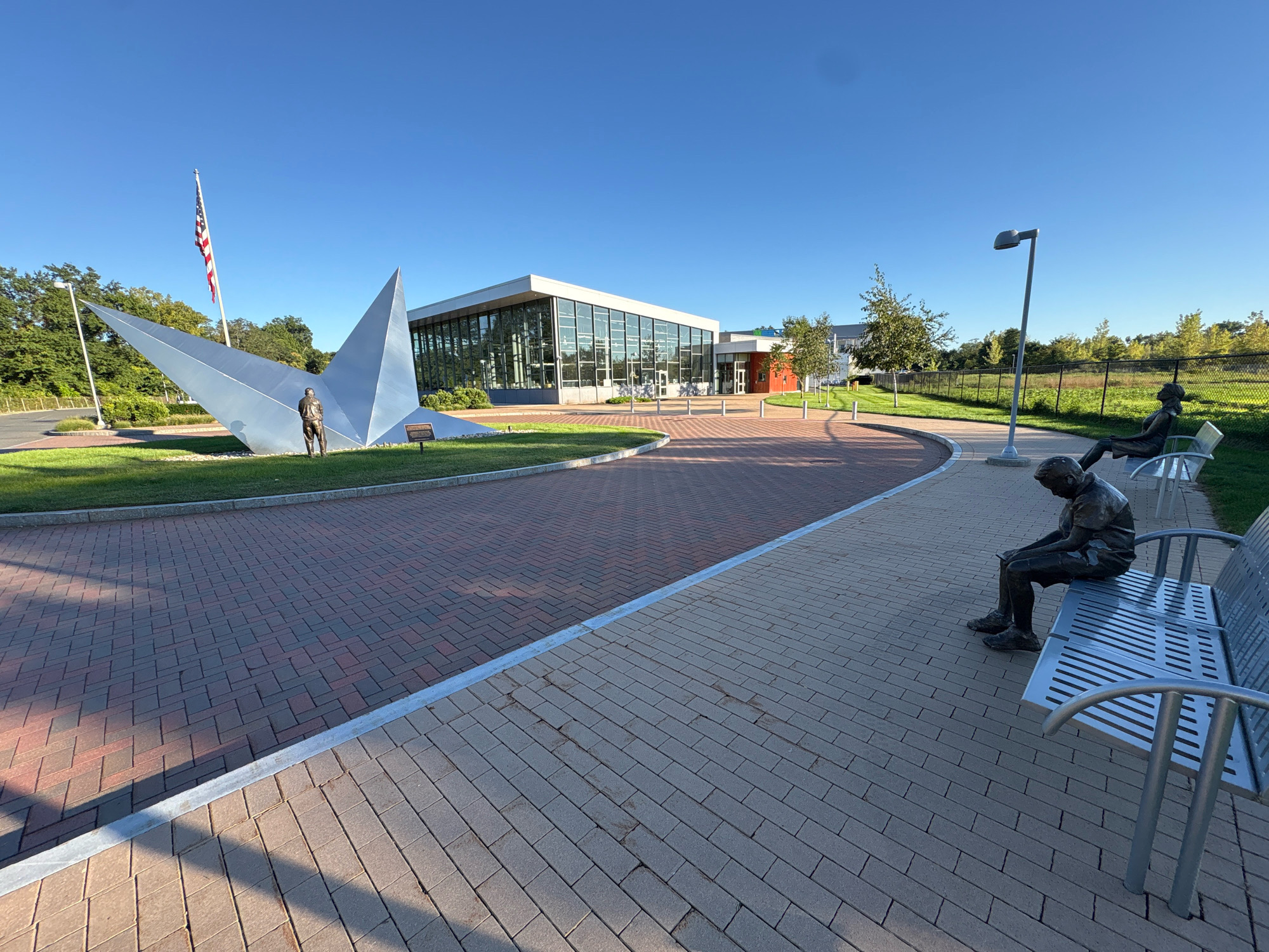 The image depicts the exterior of a modern building with a glass facade, situated in East Hartford, Connecticut, United States of America. The building is surrounded by a landscaped area with a brick-paved walkway and green spaces. A large, abstract metallic sculpture resembling a pointed star is prominent in the foreground, with a small bronze statue of a man standing near its base. An American flag flies on a flagpole behind the sculpture. To the right of the walkway, there are benches with bronze statues of seated figures, appearing to be in contemplative poses. A street lamp is visible near one of the benches. The sky is clear blue, indicating a sunny day. The scene suggests a public space, possibly a park or an entrance to a civic building.