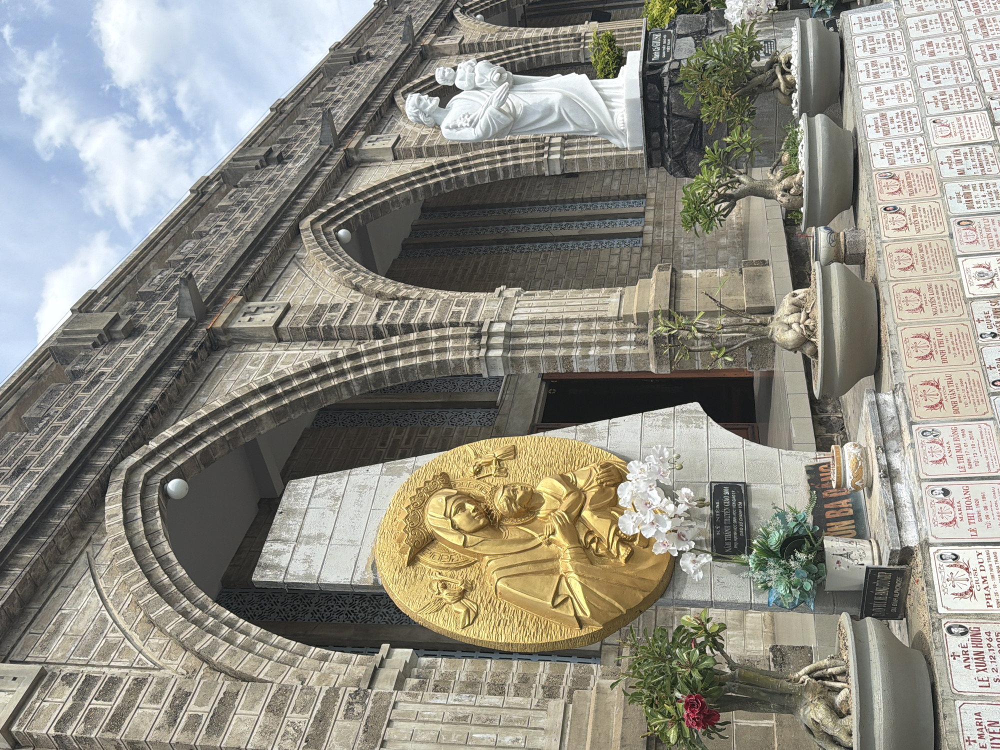 A wide-angle, eye-level shot captures the facade of a stone church in Nha Trang, Vietnam. The architecture features multiple arched entrances, characteristic of Gothic Revival style, with weathered stone construction. To the left of the central entrance, a large, circular, golden relief depicts the Virgin Mary holding the infant Jesus, adorned with a crown and surrounded by cherubs. In front of this relief, potted plants, including bonsai trees with gnarled roots, and a bouquet of white orchids are arranged. To the right of the central entrance, a white marble statue of Saint Joseph holding the infant Jesus stands on a stone pedestal. The foreground displays a row of gravestones or memorial plaques, each bearing names and dates, suggesting a cemetery or memorial garden associated with the church. The sky above is a mix of blue with scattered white clouds. The overall scene is one of religious reverence and remembrance.