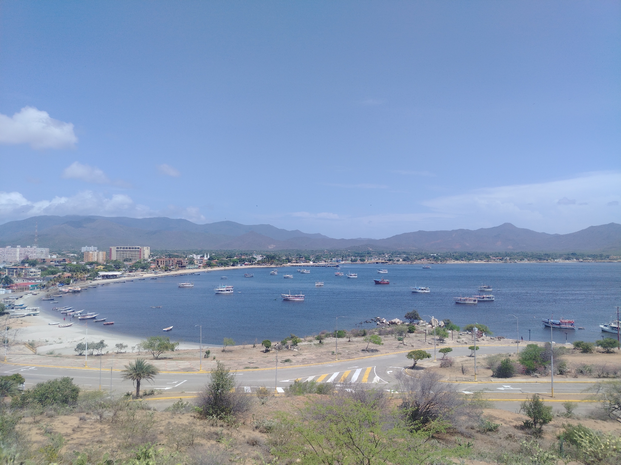This is a wide-angle, aerial view photograph of a bay in Juan Griego, Venezuela, under a clear blue sky with scattered clouds. The scene depicts a coastal town with buildings lining the shoreline, a calm blue body of water filled with numerous boats of various sizes, and a semi-arid landscape in the foreground.
In the mid-ground, the bay curves from the left to the right, showcasing a beach area where several smaller boats are pulled ashore. A paved road with yellow and white markings runs parallel to the bay, with a pedestrian crossing. A single palm tree stands near the road. The water is populated by many moored and moving vessels, including larger passenger boats and smaller fishing boats. Some of these boats have distinct red and white hulls.
The background features a range of mountains that form a natural backdrop to the town. The town itself is characterized by low-rise buildings, including apartments and commercial structures, extending along the coast. The vegetation in the foreground is sparse, typical of a dry climate, with scattered bushes, shrubs, and cacti. The overall impression is a bright, sunny day in a coastal Venezuelan town. No people are clearly discernible in the image.