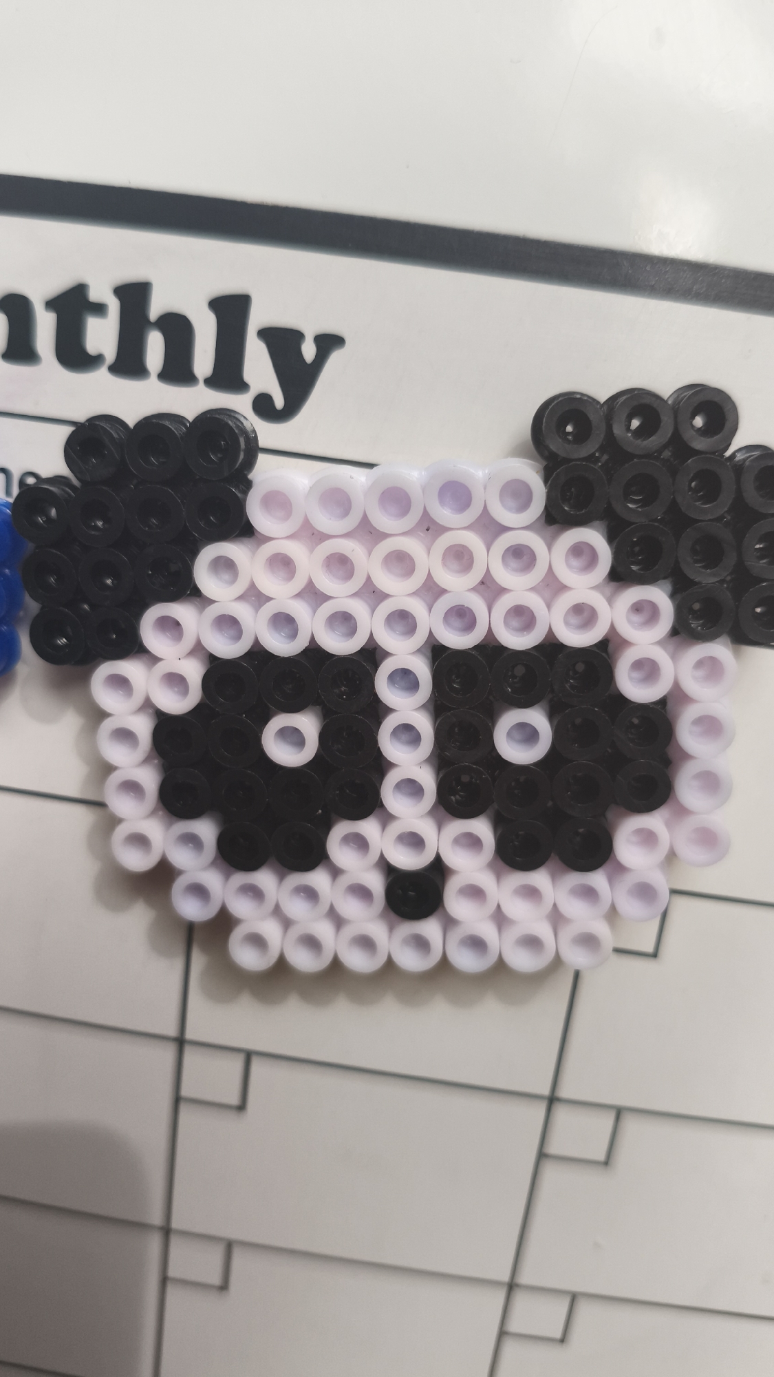 The image depicts a panda face crafted from black and white Perler beads. The panda has black ears and eye patches, surrounded by white beads. The background includes a portion of a whiteboard calendar, with the word "monthly" visible at the top left. Grid lines of the calendar are also noticeable. The location in Nowy Tomyśl, Poland cannot be determined from the image.