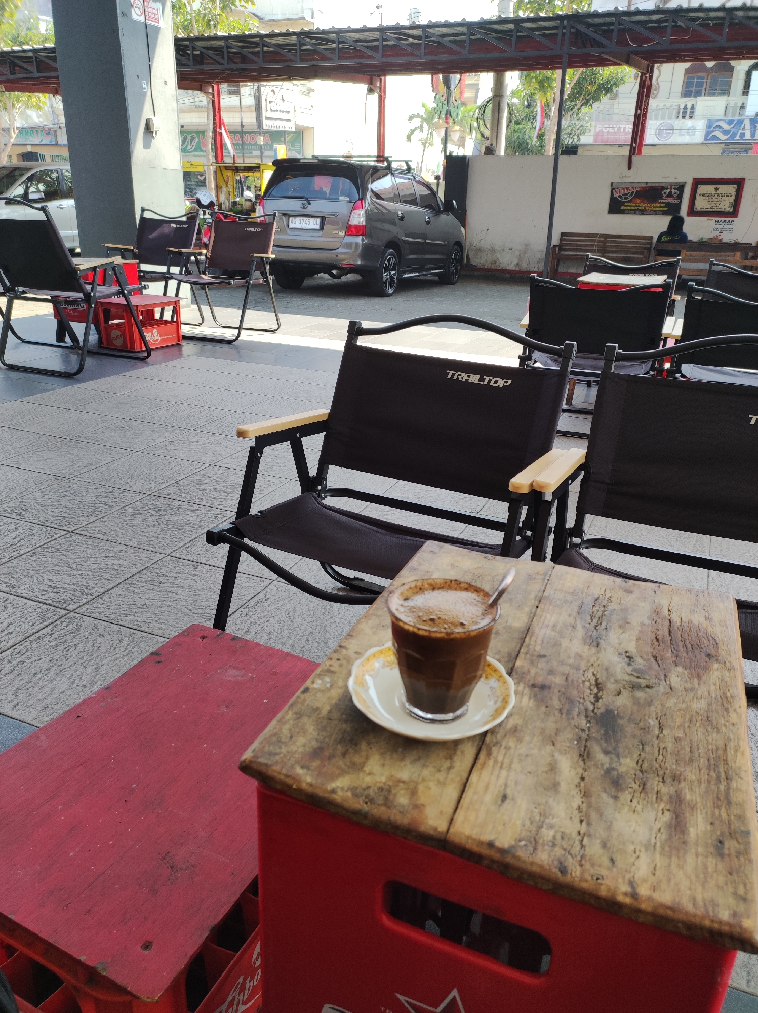 The image depicts an outdoor seating area, likely a cafe or roadside eatery, in the city of Blitar, Indonesia. In the foreground, a small wooden table, constructed from weathered planks and resting atop a red plastic crate, holds a glass of dark coffee with froth and a spoon. Beside it, another red crate is partially visible. Behind this setup, several dark folding chairs with "TRAILTOP" branding are arranged on a tiled floor. In the midground, a silver car with license plate "AG 1745 DL" is parked. The background reveals a covered walkway with supporting pillars, a building with advertisements, and other seating areas. Sunlight filters through the overhead structure, casting shadows and illuminating the scene. No individuals are clearly visible interacting with the coffee or chairs. The environment appears to be an open-air commercial space.