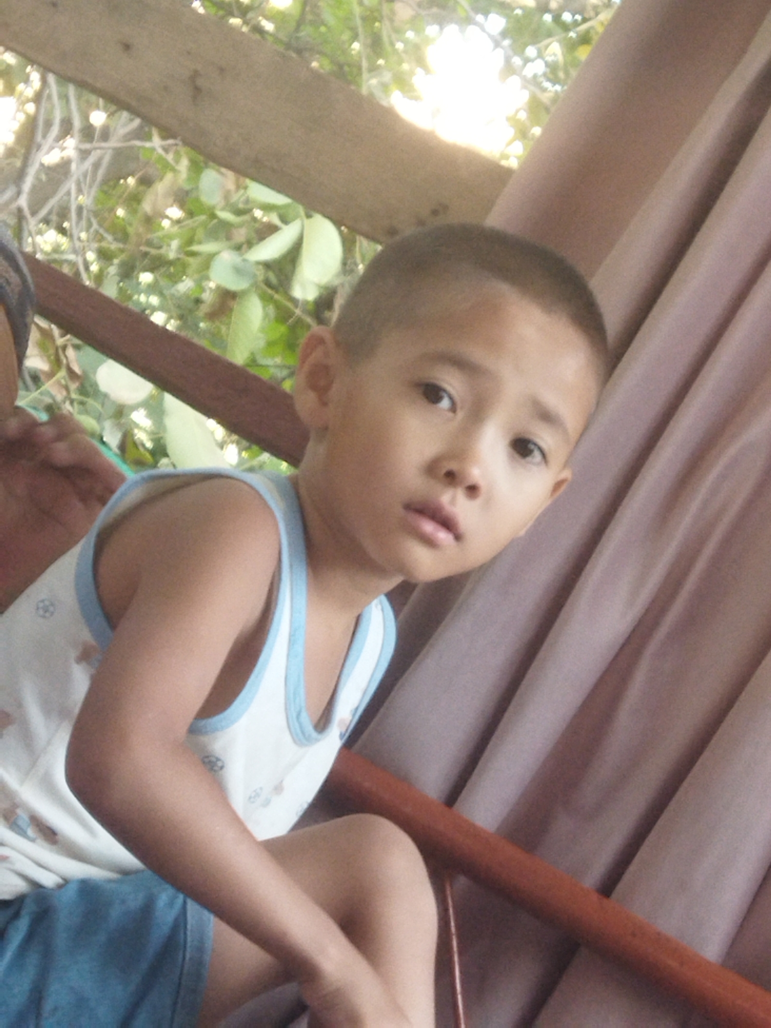 The media file shows a close-up of a young boy, likely of East Asian descent, sitting outdoors. He has short dark hair and is wearing a white tank top with blue trim and blue shorts. His expression is neutral, with his eyes looking slightly to the left. Behind him, there is a blurry background of green foliage and branches, suggesting a natural setting. To his right, there is a pleated, dusty rose-colored fabric, possibly a curtain or drapery. A wooden beam is visible at the top of the frame, and a reddish-brown wooden pole runs diagonally across the lower half of the image. The lighting suggests it is daytime, possibly with bright sunlight filtering through the trees. There is no visible text in the image. The context provided places this scene in Mailuu-Suu City, Kyrgyzstan.