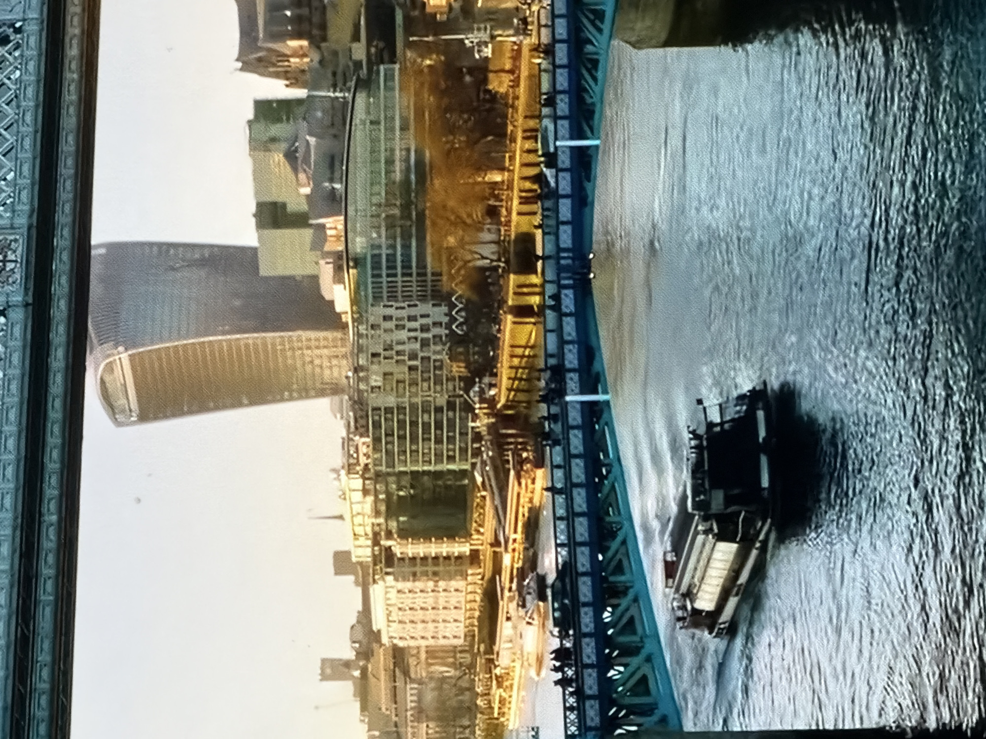 The image depicts a vibrant urban waterscape under a bright, warm light, likely late afternoon or early morning. The scene is framed at the top and sides by a dark, indeterminate structure, suggesting a view from beneath another bridge or an opening.
In the mid-ground, a teal-blue metal bridge, identified as Southwark Bridge, spans a wide river, the River Thames. The bridge features intricate metalwork and a pedestrian walkway on both sides. Numerous people are visible walking across the bridge. A dark vehicle, possibly a car, is also present on the bridge deck.
Beneath the bridge, a long, dark passenger boat with illuminated windows traverses the river from left to right, creating a distinct wake. The river water is rippled and reflects the warm sunlight. Further upstream on the left, another smaller, lighter-colored boat is partially visible.
The background reveals a dense cityscape along the riverbank on the northern side. The most prominent structure is the distinctive "Walkie-Talkie" skyscraper (20 Fenchurch Street), with its curved, tapering profile, situated towards the center-right. Adjacent to it and further left, other modern glass-fronted buildings are visible, including the partial view of another iconic structure (likely the Gherkin) further to the left of the Walkie-Talkie. A mix of contemporary and older brick buildings lines the river, all bathed in the golden light. Trees with bare branches are interspersed along the riverbank, suggesting a temperate season. The sky above is bright and clear, indicating a sunny day.
The depicted location is the city of London, United Kingdom.