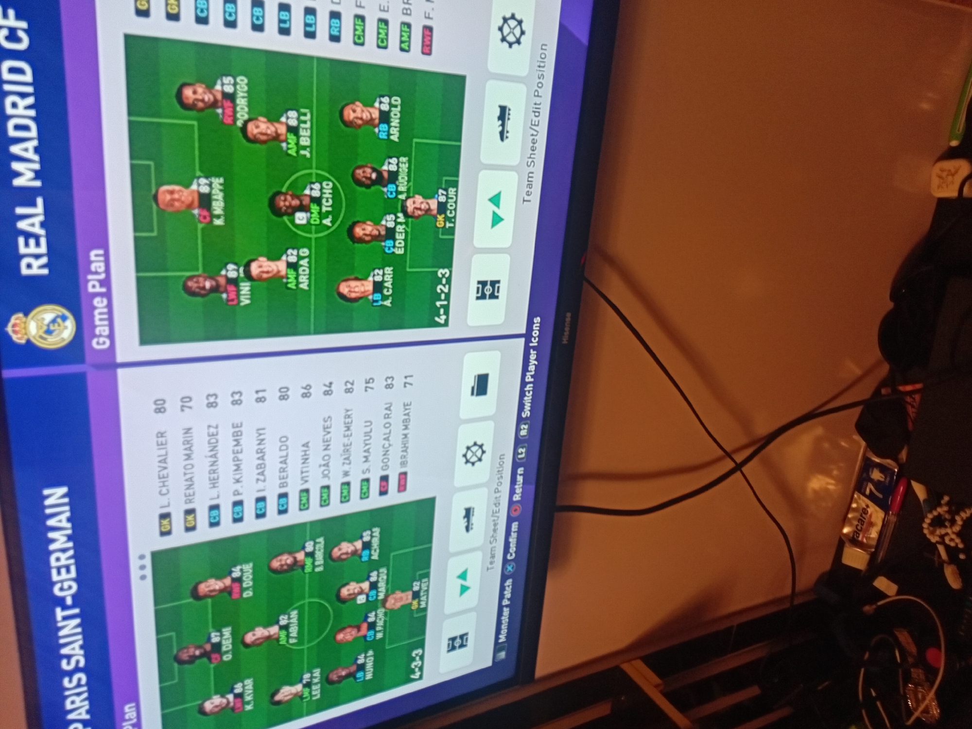 A vertically oriented photograph captures a TV screen displaying a football video game's team management interface. The screen is divided into two main sections, showcasing the "Game Plan" for two different clubs: Paris Saint-Germain (PSG) on the left and Real Madrid C.F. on the right.
PSG's formation is a 4-3-3, with players like K. KYAR (LMF 86), LEE KAI (LMF 78), O. DEME (LWF 87), D. DOUE (RWF 84), FABIAN (AMF 82), B. BARCOLA (DMF 86), NUNO (LB 84), W. PACHO (CB 84), MARQUI (CB 84), ACHRAF (RB 85), and MATVEI (GK 82) on the field. A list of substitute players with their ratings and positions is also visible, including L. CHEVALIER (GK 80), RENATO MARIN (GK 70), L. HERNÁNDEZ (CB 83), P. KIMPEMBE (CB 83), and others.
Real Madrid's formation is a 4-1-2-3, featuring players such as VINI (LWF 89), K. MBAPPE (CF 89), RODRYGO (RWF 85), ARDA G (AMF 82), J. BELLI (AMF 88), A. TCHO (DMF 86), A. CARR (LB 82), ÉDER M (CB 85), A. RÜDIGER (CB 86), ARNOLD (RB 86), and T. COUR (GK 87).
Below the formations, various game interface options are visible, including "Team Sheet/Edit Position," "Monster Patch," "Confirm," "Return (L2)," and "Switch Player Icons (R2)." A "Hisense" logo is faintly visible on the bottom right bezel of the TV.
The TV screen is positioned against a plain, light brown or beige wall. Cables are draped on the wall and a surface to the right of the screen. Partially visible on this surface are a small white package labeled "care+" and "7", a red object, and some miscellaneous dark items, possibly indicating a bedside table or shelf. The lighting in the room appears dim, suggesting an indoor evening setting.