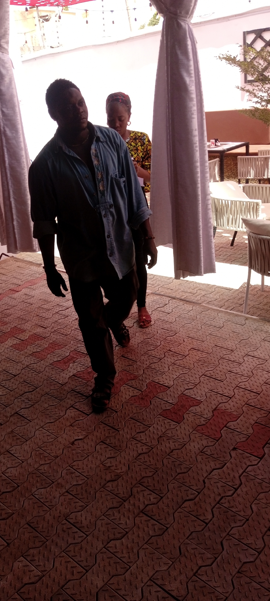 The image captures a scene in Sabon Gari East, Nigeria. A man in a blue denim shirt and dark pants is walking towards the camera on a tiled floor with a geometric pattern. Behind him, a woman wearing a patterned dress and head covering follows. The background shows an outdoor seating area with white chairs and tables, partially obscured by white curtains. A red awning is visible overhead.
