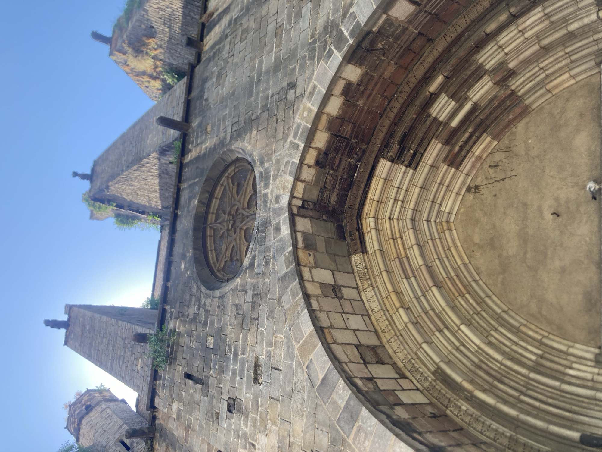 The image shows a close-up, low-angle view of an ancient stone structure, possibly a church or cathedral, against a clear blue sky. The perspective is tilted, making the structure appear to loom upwards. The stonework is rough-hewn and weathered, with visible age and texture.
Dominating the right side of the frame is a large, arched opening, elaborately decorated with multiple concentric rings of carved stone. The inner rings are a lighter color and appear smoother, while the outer rings are darker and show more intricate patterns. The archway leads into a dark, indistinct interior.
To the left of the archway, on the main wall of the building, is a circular window with delicate tracery, resembling a rose window. This window is also made of stone and has a complex geometric pattern. The wall itself is constructed from numerous rectangular stone blocks, some of which are irregular in shape and size, suggesting a long construction period or significant repairs over time.
In the upper left and lower left of the image, parts of tall, pointed towers or buttresses are visible. These also appear to be made of stone and are topped with architectural elements that suggest gargoyles or decorative finials. Some greenery, likely moss or small plants, grows from the crevices and tops of these structures, indicating that the building is exposed to the elements and has been standing for a considerable time.
The sunlight suggests it is daytime, likely morning or afternoon, casting soft shadows and highlighting the textures of the stone. The clear sky implies fair weather. The overall impression is one of historical grandeur and enduring architecture. The location is identified as Barcelona, Spain, which is consistent with the Gothic architectural style often found in the city. There is no visible text in the image.