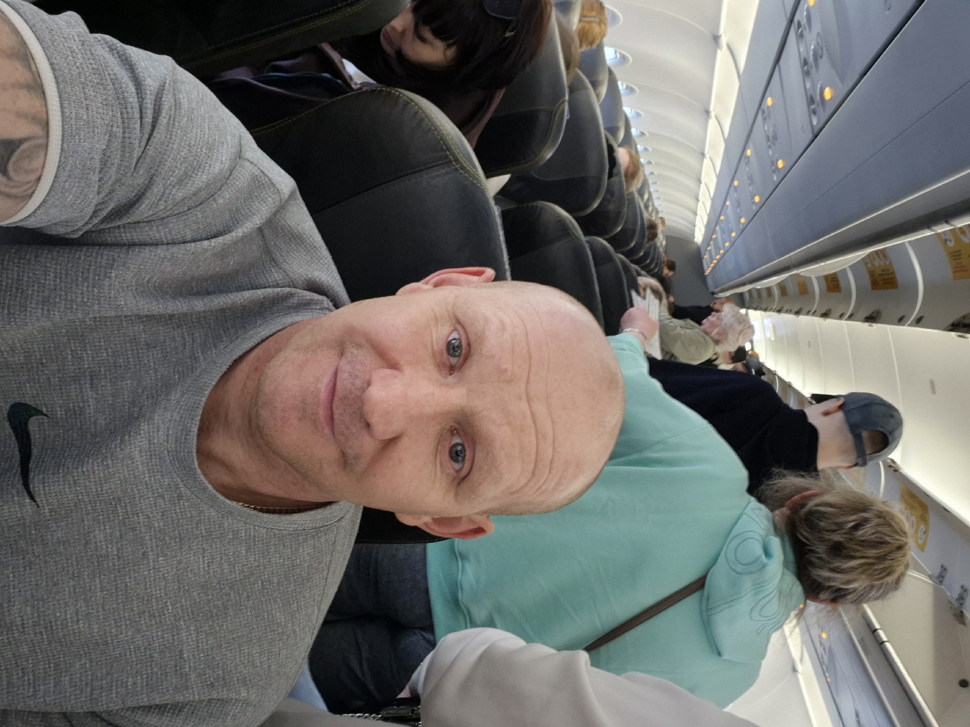 The image is taken inside an airplane cabin. A bald man with blue eyes and a grey t-shirt takes a selfie. He has a tattoo on his arm. Behind him are other passengers in their seats. A woman with dark hair is sleeping, and a woman with blonde hair is seated in front. Overhead compartments and passenger controls are visible along the plane's interior. The image does not depict the city of Ufa, Russia.