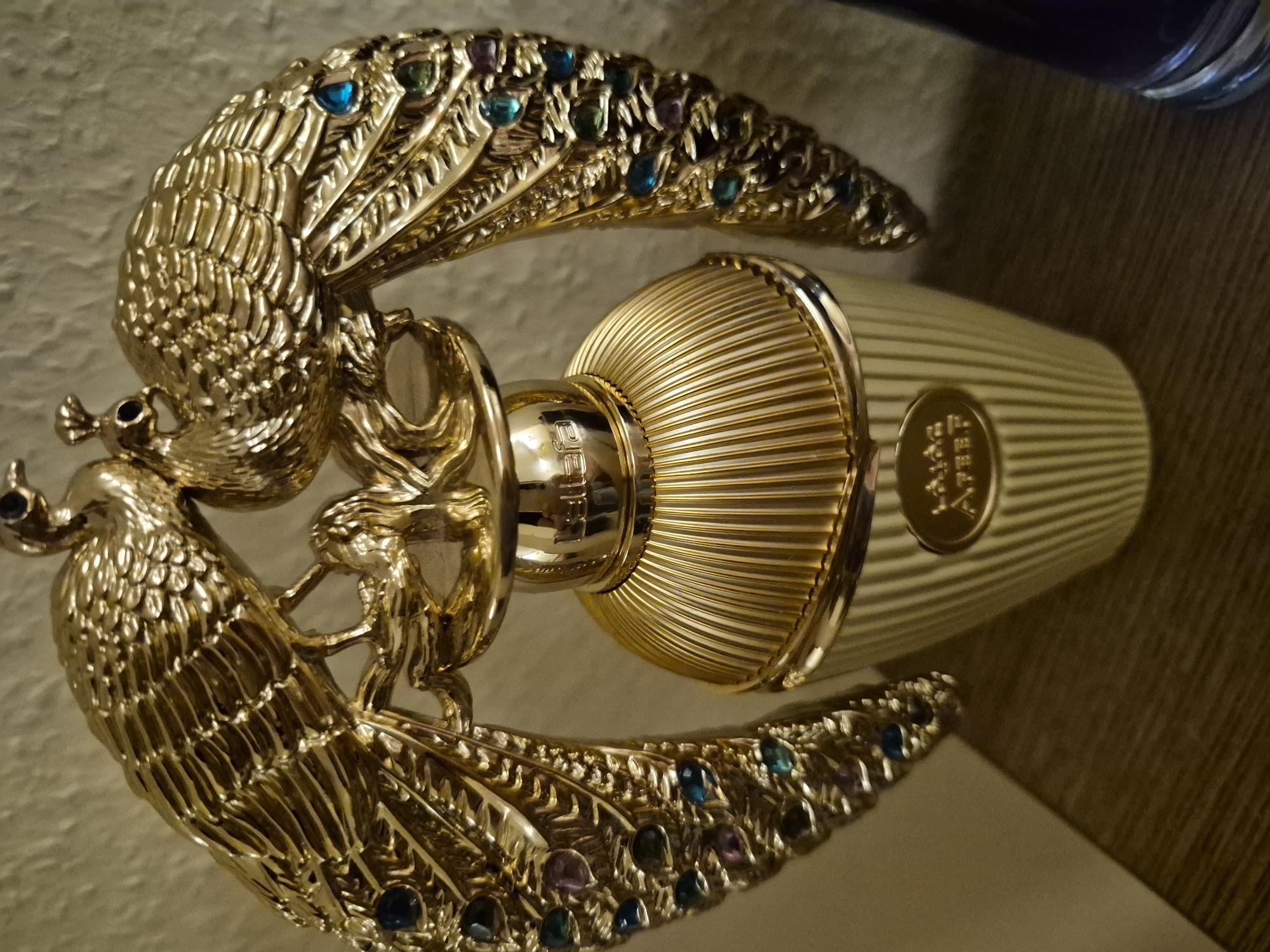 A close-up, eye-level, indoor shot features an ornate, gold-colored perfume bottle with two decorative peacock figures adorning its top. The bottle itself is ribbed and stands on a surface that appears to be a shelf made of wood with a dark grain. The background is a textured, light-colored wall. The peacock figures, also gold, have outstretched wings embellished with colorful gemstones that appear to be blue, purple, and green. The light source casts subtle shadows, highlighting the metallic sheen of the peacocks and the ribbed texture of the bottle. The bottle has a golden cap and a raised, oval emblem on its body containing Arabic script. The overall impression is one of luxury and intricate craftsmanship.