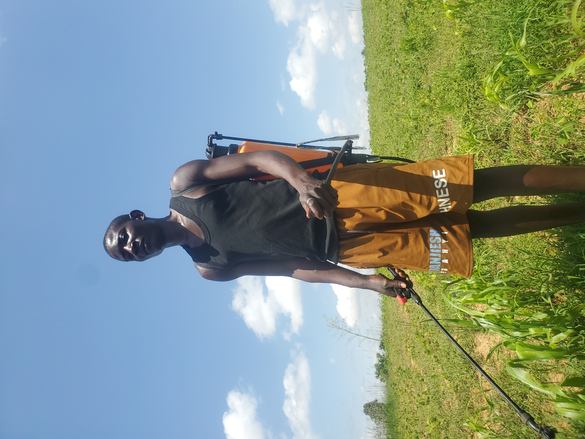 A dark-skinned man stands in a field under a bright blue sky with scattered clouds, wearing a black tank top and brown shorts. He is holding a sprayer with an orange tank on his back and a long wand in his right hand. The field is covered in green vegetation, with some taller stalks of corn in the foreground. The man appears to be looking up and to the left. The image was taken in Wumat Central, Nigeria.