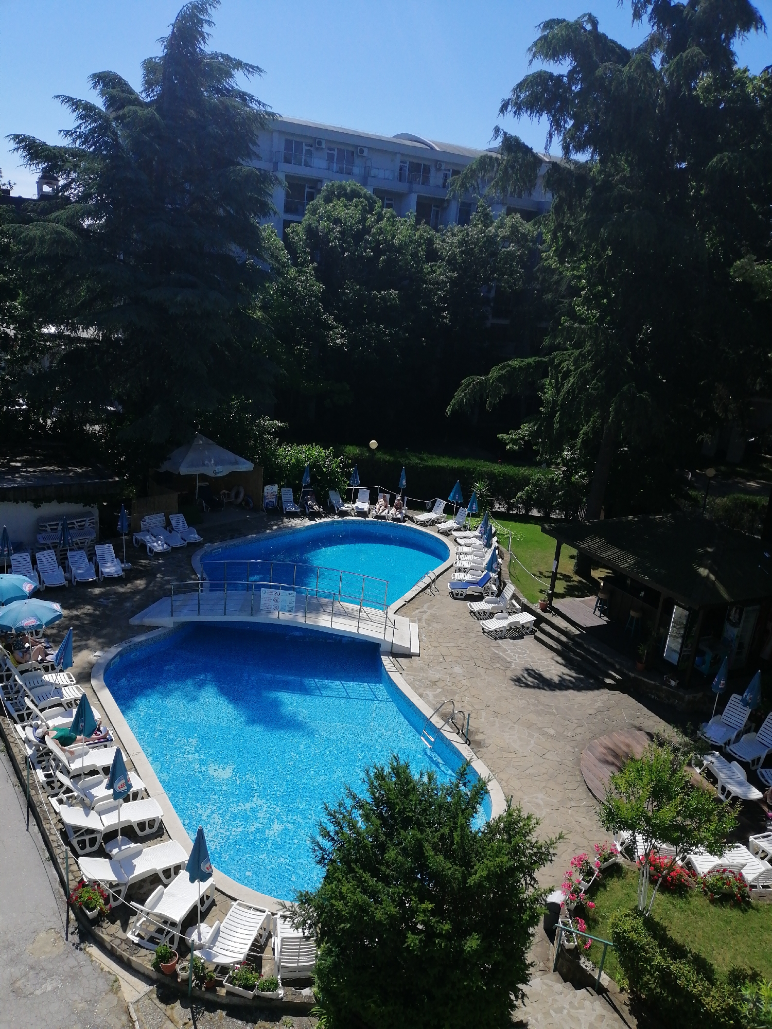 The image depicts an outdoor swimming pool area at a resort or hotel on a sunny day. The scene is dominated by two large, irregularly shaped swimming pools filled with bright blue, tiled water. A small, white pedestrian bridge with railings spans across a section connecting the two pools.
Surrounding the pools are numerous white lounge chairs, many of which are occupied by people relaxing or sunbathing. Several blue and green beach umbrellas provide shade. To the left of the pools, a paved pathway leads to more lounge chairs and a wall. In the background, a multi-story building with a light-colored facade and numerous windows is visible, partially obscured by dense, mature trees. A small, open-air bar or kiosk with a wooden roof and seating is situated to the right of the pools, on a grassy area.
The weather appears to be clear and sunny, indicated by the bright lighting and the clear blue sky. The time of day is likely midday or early afternoon, given the strong shadows cast by the trees and structures. The presence of lounge chairs, umbrellas, and swimming pools strongly suggests a recreational setting, possibly a vacation destination. The lush greenery and the building in the background hint at a location in a resort town.
There are some visible signs with text, but they are too small and blurry to be read. One sign is visible on the bridge, and others are placed near the lounge chairs and at the bar area. The overall mood of the image is one of relaxation and leisure.