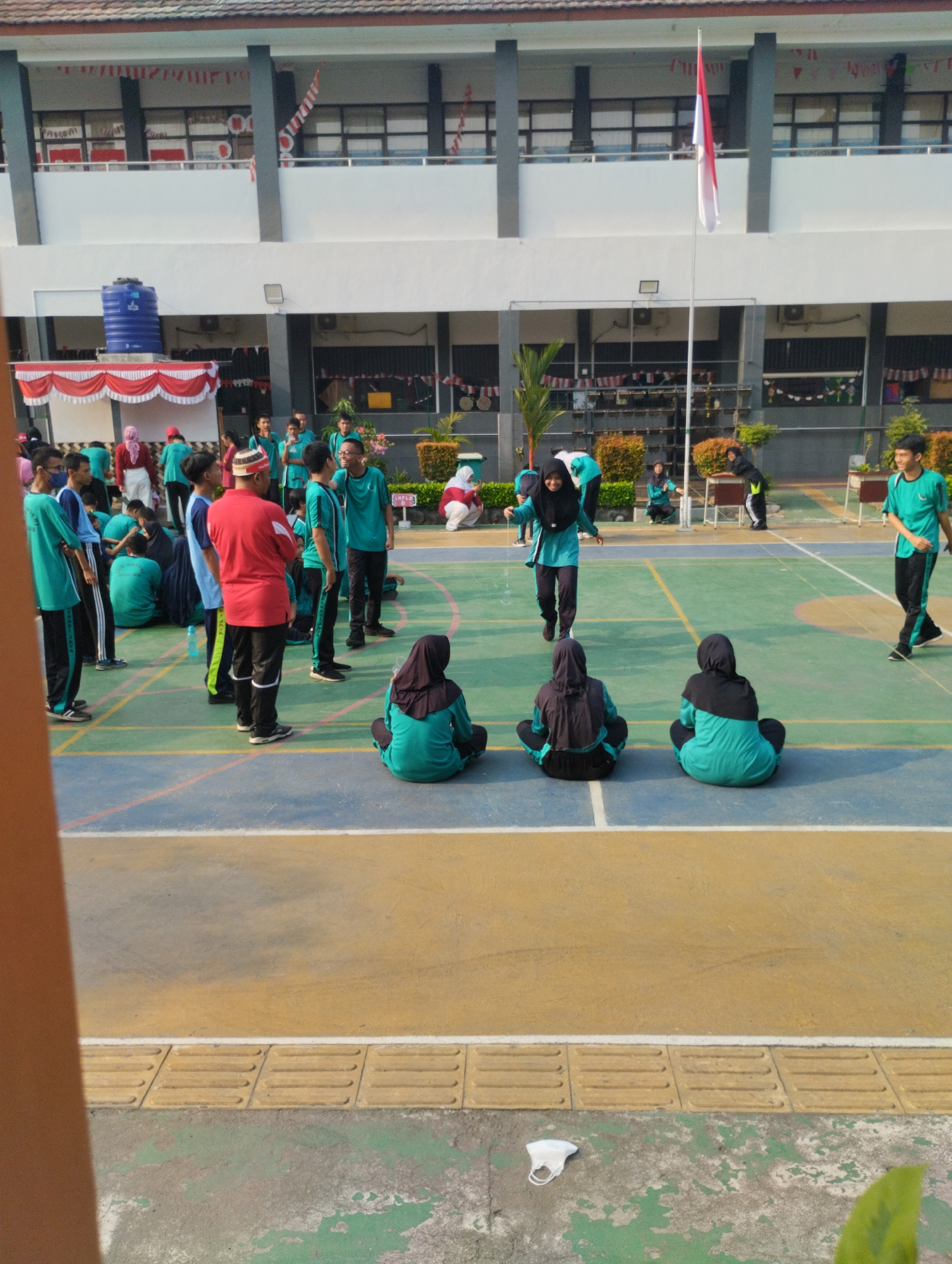 The image captures a scene on an outdoor basketball court in front of a multi-story building. Several individuals, primarily young people wearing teal athletic uniforms with black pants, are present on the court. Some are standing, engaged in conversation, while others are seated. One person in a teal hijab is in motion, appearing to jump or dance, with a slight blur indicating movement. Another individual in a teal hijab is also seated on the court. In the background, a building with white and gray sections is visible, adorned with red and white Indonesian flags and pennants. A blue water tank is also situated near the building. The court itself is marked with lines in yellow and blue. In the foreground, a discarded white face mask lies on the ground. The overall impression is a school or sports event taking place outdoors. The specific location in the city of Bekasi, Indonesia, is suggested by the presence of Indonesian flags.