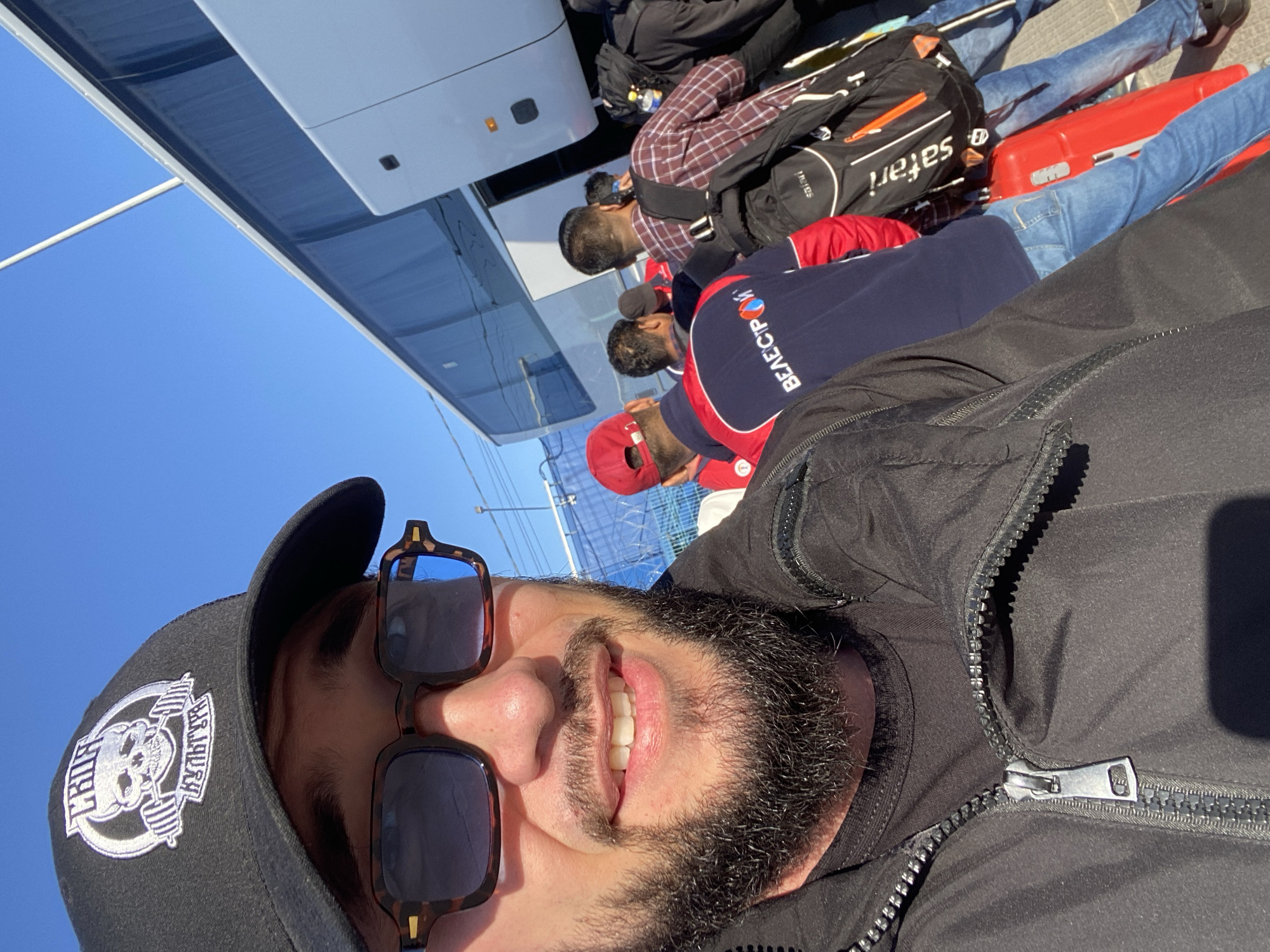 In Koskolovo, Russia, under a clear blue sky, a man wearing a black baseball cap with a skull logo and sunglasses smiles at the camera. He is dressed in a black jacket over a dark t-shirt, with the zipper partially open. Behind him, a group of people are queuing up to board a white bus. Some individuals are carrying backpacks, and one person has a bright red suitcase. The bus has tinted windows and its door is open, indicating people are in the process of boarding. The scene appears to be a departure or arrival point, likely for a group tour or transport.