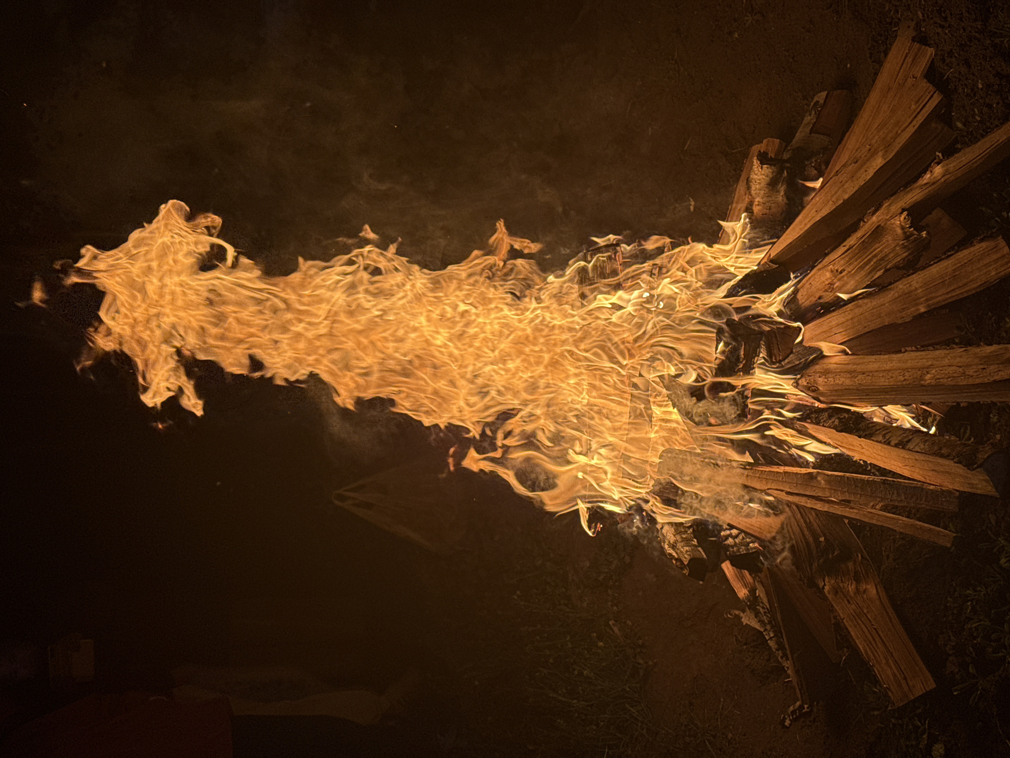 The image shows a bonfire at night. The fire is burning intensely, with flames extending horizontally to the left. The fuel for the fire appears to be a pile of wood, arranged somewhat haphazardly on the right side of the image. The surrounding area is dark, suggesting the fire is the primary light source. There are hints of ground and vegetation in the lower right. There is no information about the location Ust-Koksa, Russia in the image.
