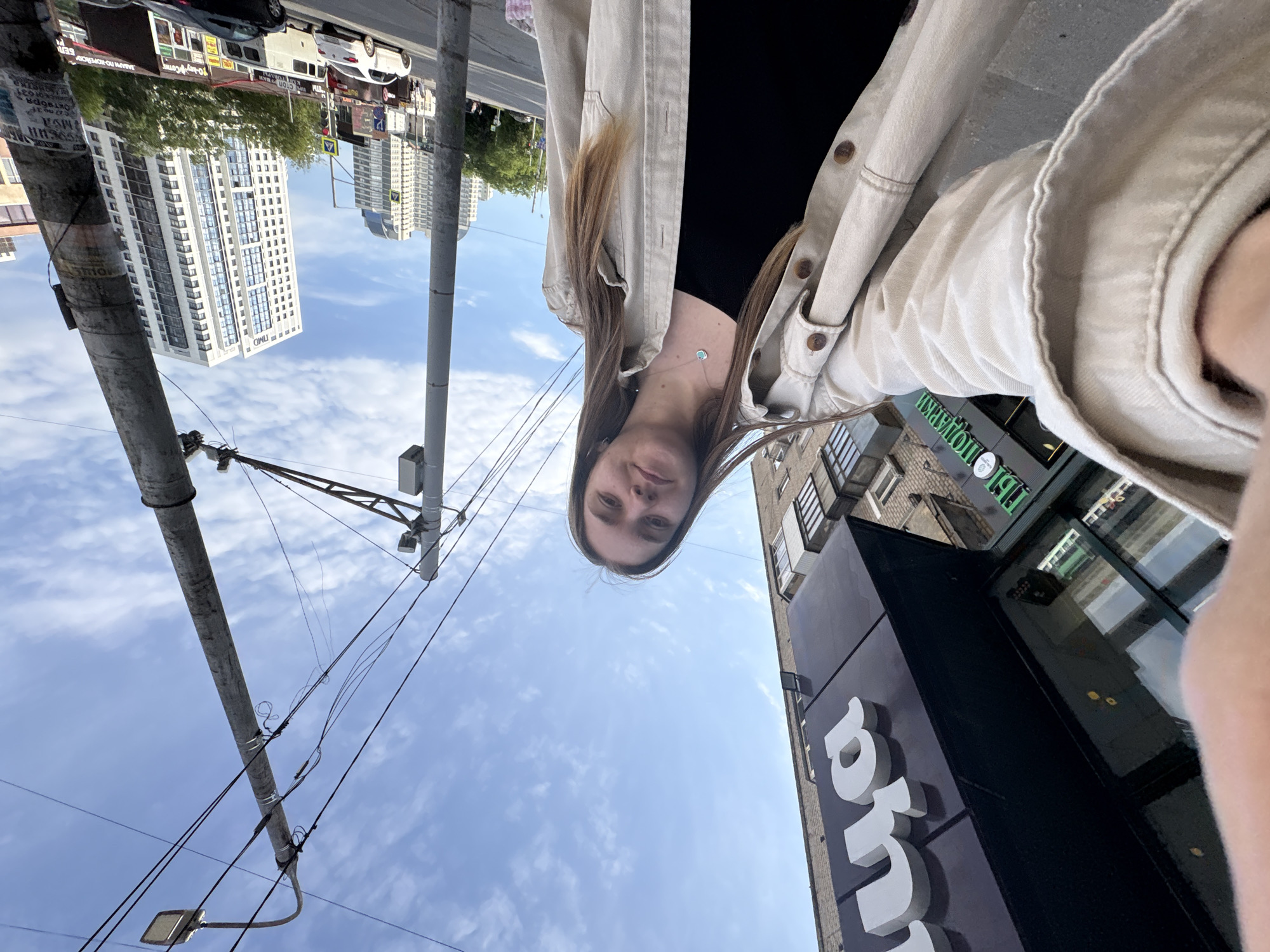 A vertically oriented, ground-level selfie shows a young woman in Perm, Russia, facing down at the camera. She wears a black top under a beige, button-down jacket. Her long brown hair hangs down, and a delicate necklace is visible.
Behind her, the Perm city landscape is upside down. Modern, high-rise apartment buildings with white facades are seen in the distance, along with various commercial buildings. Utility poles with overhead wires are positioned against the blue sky and scattered clouds. Part of a store with large white letters that seem to spell "Поляна" in Cyrillic is visible on the right.
