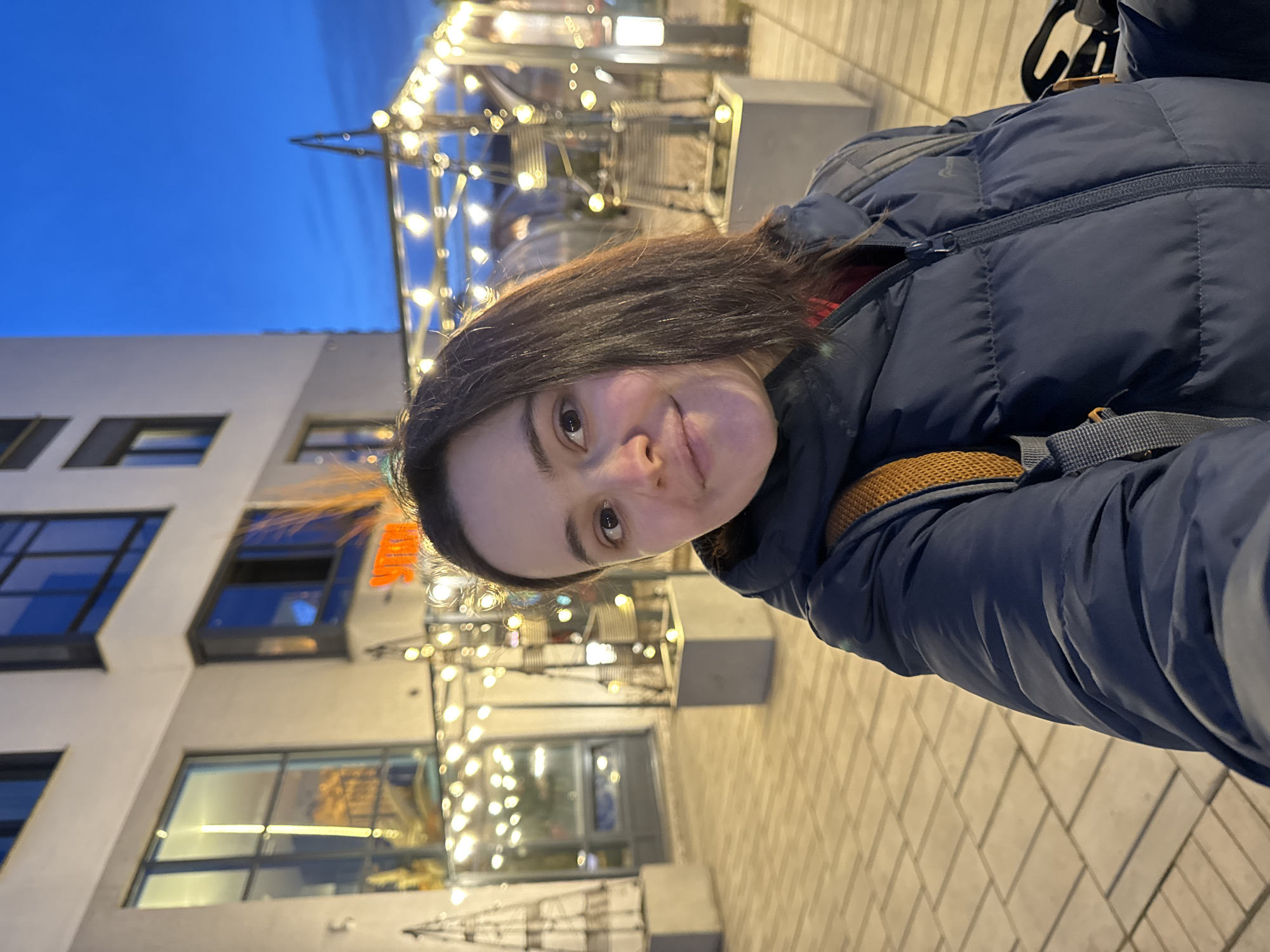 A woman is standing on a brick-paved area next to a building with windows. String lights are hung overhead. She wears a dark blue puffer jacket and backpack. The building has modern architecture. The location is Izhevsk, Russia.