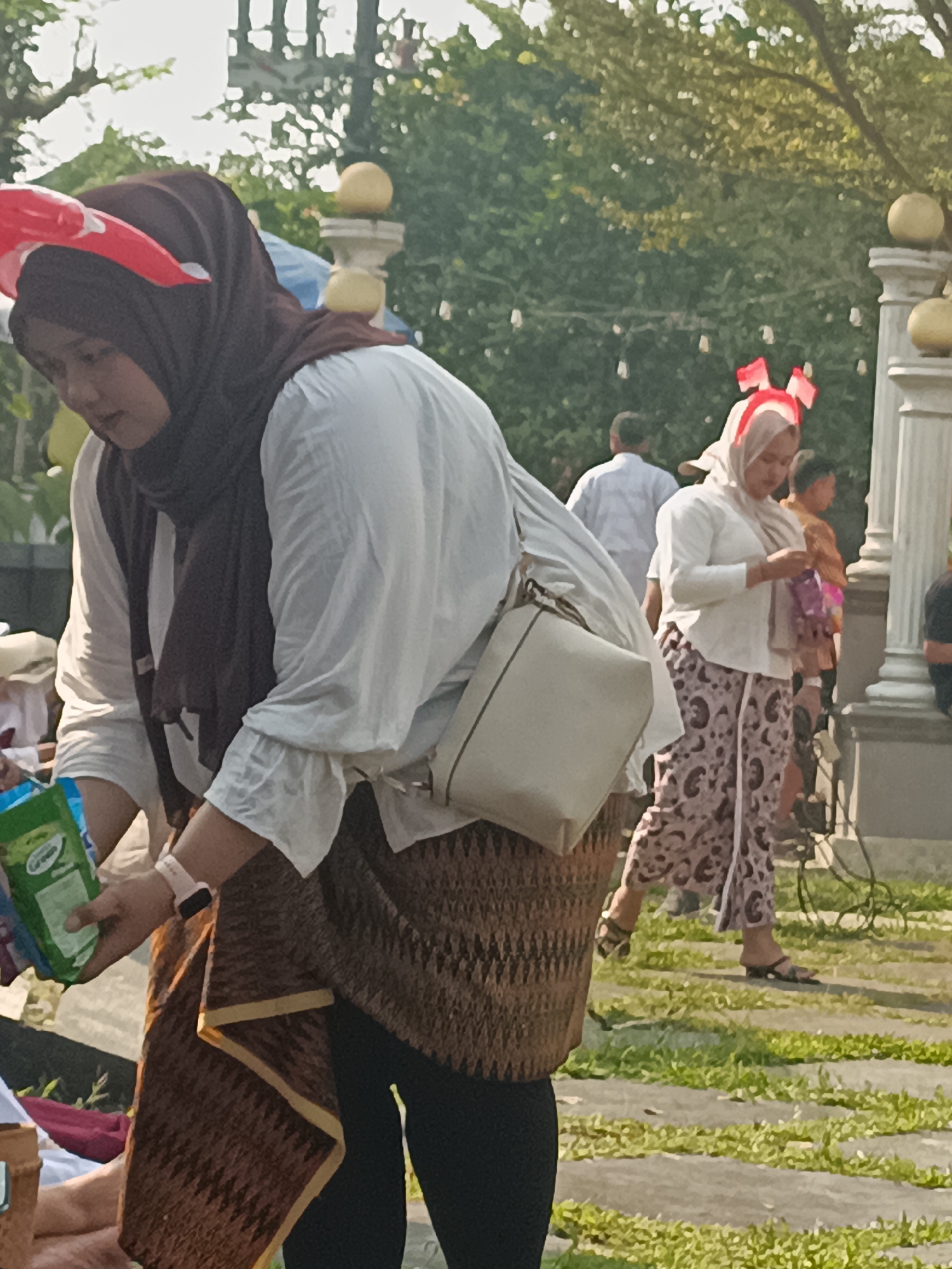 In Sleman, Indonesia, a group of people are gathered outdoors in what appears to be a park or garden setting. The foreground is dominated by a woman wearing a dark brown hijab and a white blouse. She is bent over, holding a green package in her left hand and a patterned brown skirt is visible around her waist. A light-colored crossbody bag is slung over her shoulder. To her right, another woman in a light-colored hijab and a white shirt with patterned pants stands with a small purple object in her hands. Red and white patriotic headbands or decorations are visible on both women, suggesting a national celebration. In the background, other individuals are milling about, and trees with green foliage suggest a daytime outdoor event. Architectural elements resembling pillars are also present, hinting at a public space or venue.