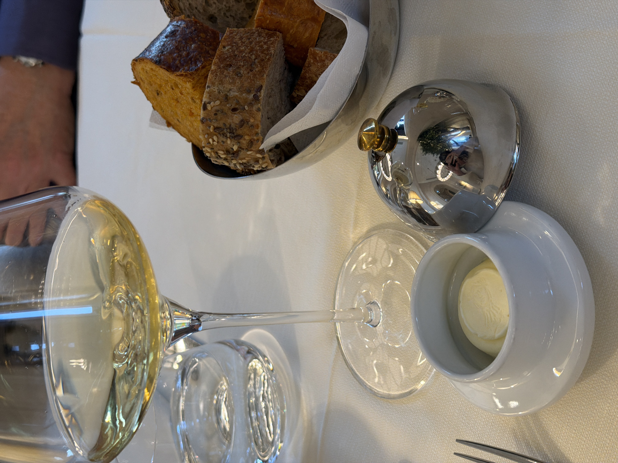 A table setting in what appears to be a restaurant in Belgrade, Serbia. A wine glass filled with white wine is partially visible, lying on its side. Next to the wine glass is another empty wine glass. A silver bread basket filled with sliced bread is present, accompanied by a covered butter dish containing butter. The scene is set on a white tablecloth. A portion of a person's arm and sleeve is visible in the upper left corner. A fork lies to the lower right. The reflection of interior features is visible in the polished surface of the butter dish cover.
