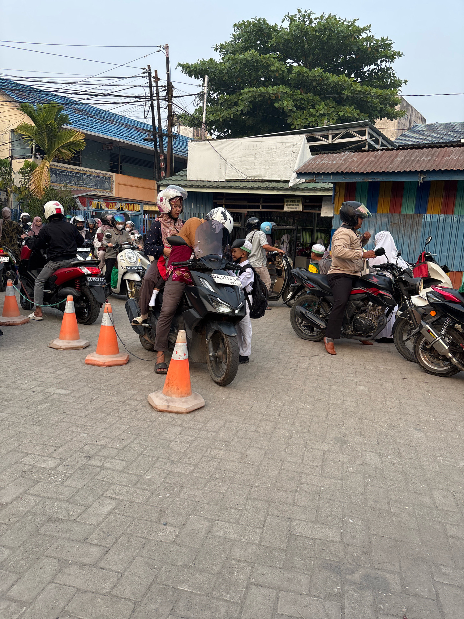 The image depicts a street scene in Banjarmasin, Indonesia, where numerous individuals are gathered around motorcycles. The foreground is dominated by a paved surface made of interlocking gray bricks. Several orange traffic cones are strategically placed on the pavement, indicating a designated area or a temporary obstacle.
A variety of motorcycles are visible, with people seated on or standing near them. Most of the people are wearing helmets, a common safety measure for motorcycle riders. Some individuals are dressed in traditional Islamic attire, such as headscarves, suggesting a cultural context. Among the crowd, there are also children, some of whom are wearing school uniforms.
The background reveals a street lined with buildings. One building has a blue roof and an orange facade with Arabic inscriptions, possibly a mosque or a religious institution. Another building features a colorful corrugated metal exterior with vertical stripes of red, green, and yellow. Overhead, a complex network of electrical wires crisscrosses the sky, supported by utility poles. Trees, including a palm tree and a large leafy tree, are interspersed among the structures, providing greenery to the urban landscape. The overall atmosphere appears to be one of daily activity, possibly related to transportation or commuting in the city.