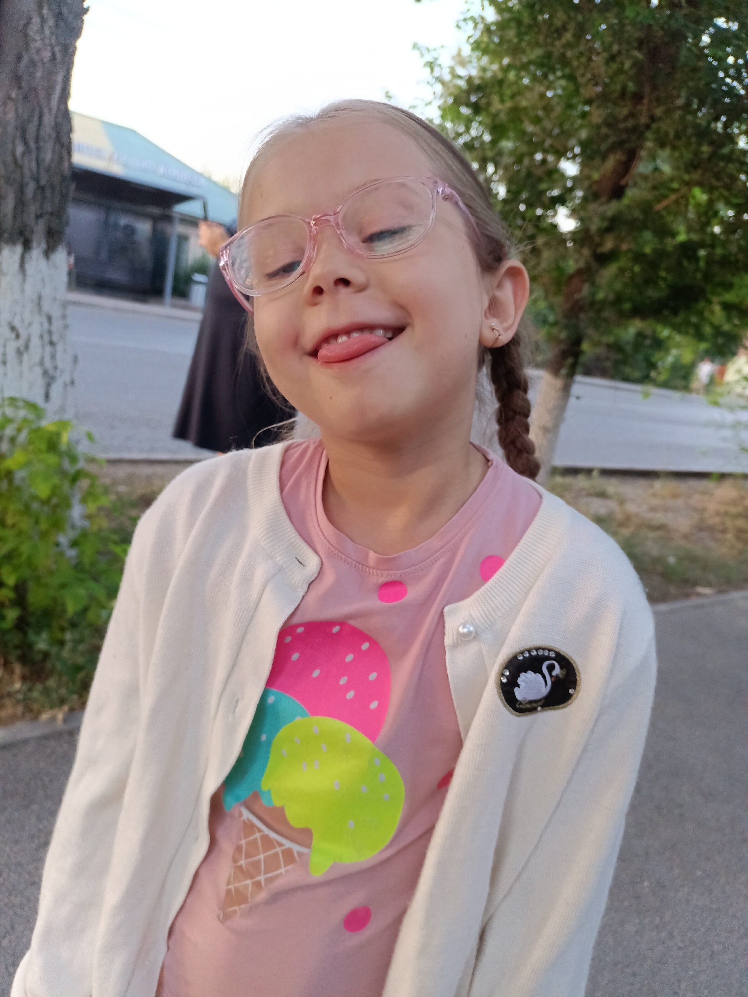 A young girl wearing pink glasses and a white cardigan is sticking her tongue out at the camera. She has blonde hair braided into two pigtails and a pink shirt with an ice cream cone design. A badge of a swan is attached to her cardigan. The background appears to be an outdoor urban setting with a building and trees. The child's expression is playful.