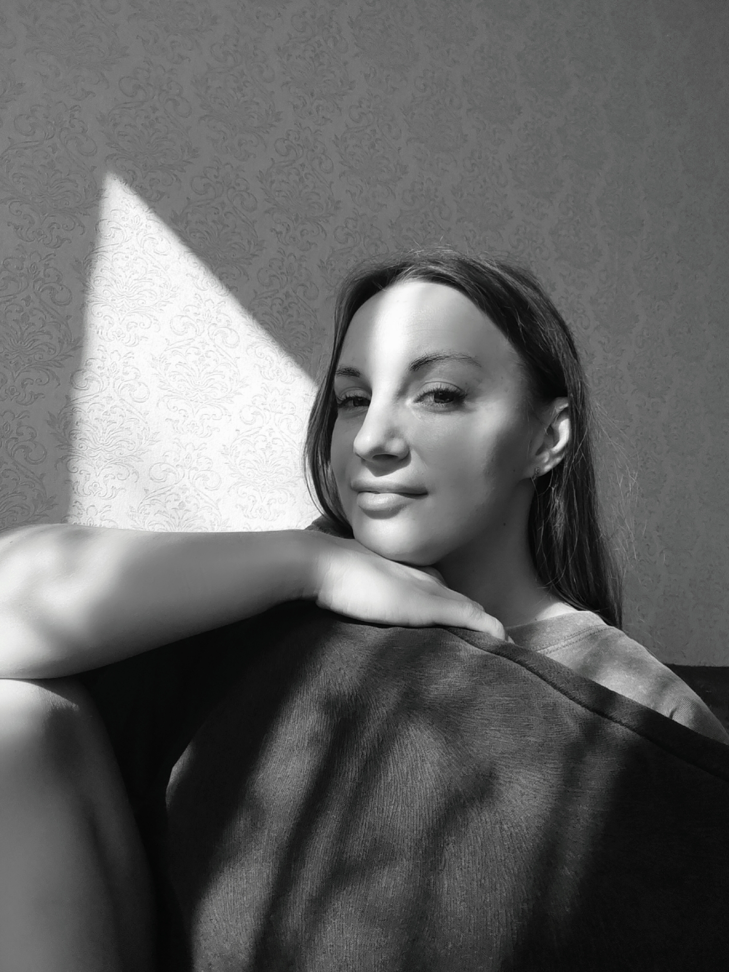 A black and white medium shot depicts a woman leaning on a dark cushion. She has long hair and is positioned in front of a wall with patterned wallpaper. A rectangular beam of light crosses the wall and her face, casting shadows. The location is unspecified, but the prompt states it is in Chaikovsky, Russia.
