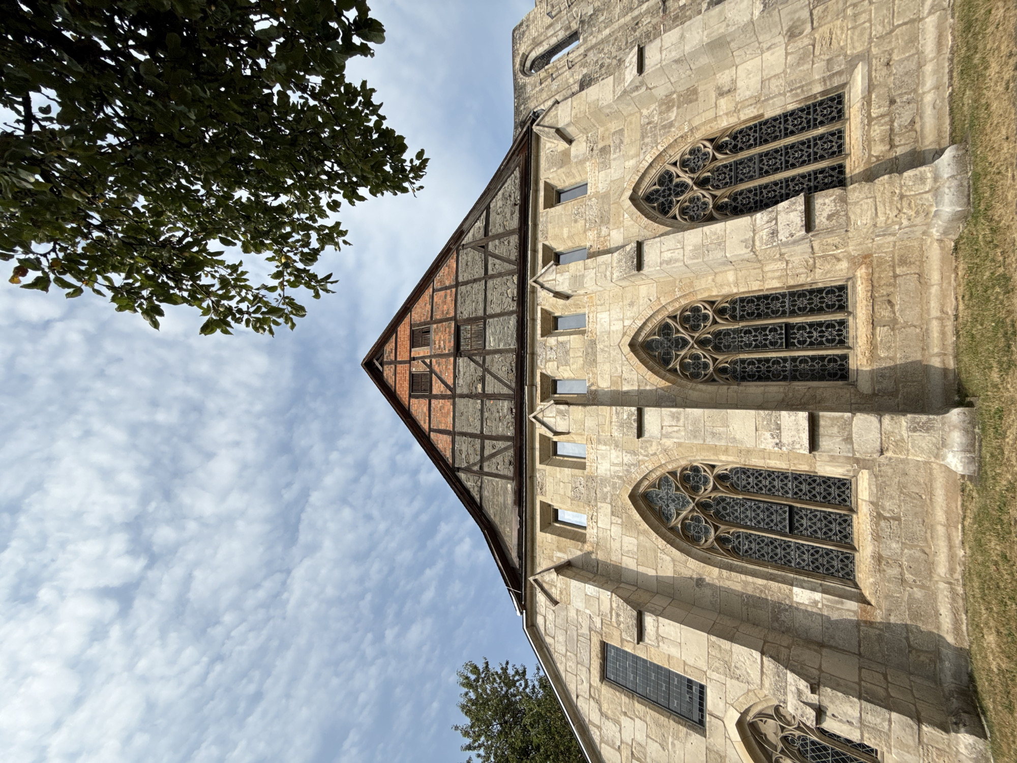 This is a low-angle, wide shot of the exterior of the Augustinian monastery in Walkenried, Germany. The stone facade of the church is visible, with several tall, arched windows featuring intricate tracery. Above the stone walls, a portion of the timber-framed gable end of the roof is visible, constructed with red brick infill. A large tree with green leaves is in the upper right corner of the frame, partially obscuring the sky. The sky is blue with scattered white, wispy clouds. The ground in front of the church is covered in dry grass. No people are present in the image.