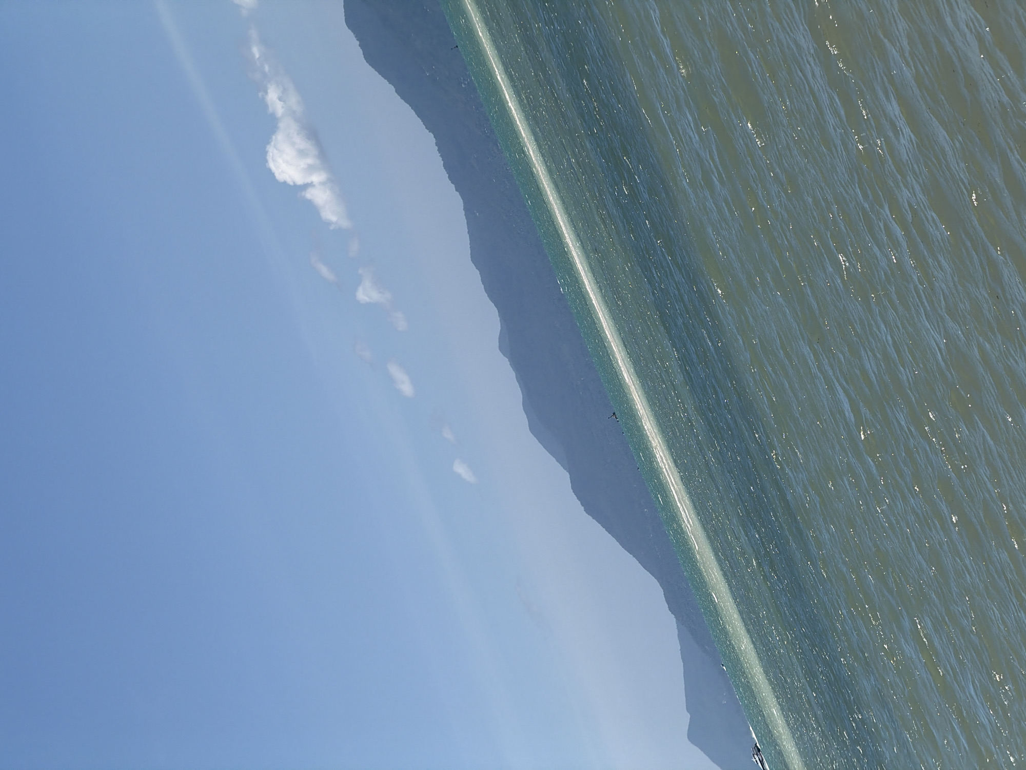 An angled, wide shot captures a bright, sunny day at sea near the coast of Ko Samui District, Thailand. The sky is a clear, vibrant blue with a few wispy white clouds. In the distance, the dark, verdant slopes of mountains rise from the water's edge, creating a silhouetted backdrop. A bright, diagonal line of sunlight reflects off the choppy, turquoise water, dividing the scene. The foreground is dominated by the textured surface of the water, with numerous small waves and glints of sunlight. A white boat is visible in the far left of the frame, moving towards the left.