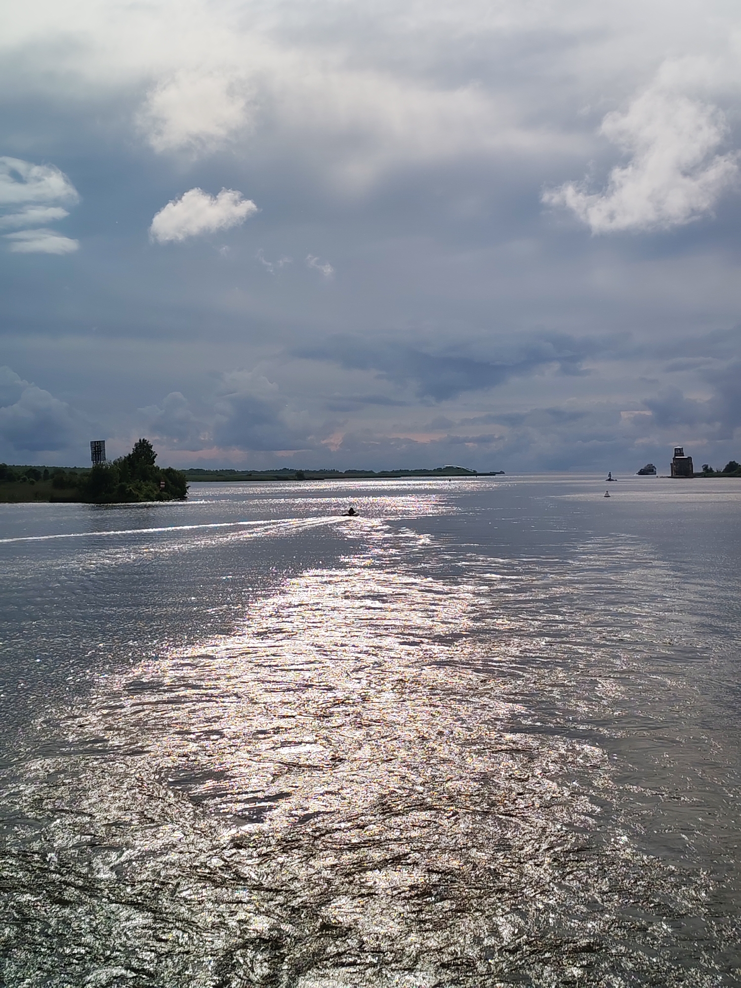 The image depicts a wide body of water under a dramatic sky with dark, heavy clouds interspersed with lighter patches. Sunlight breaks through in streaks, creating a shimmering, reflective surface on the water. In the distance, a landmass with trees and structures is visible on the left. A small boat or personal watercraft is seen moving on the water, leaving a wake that contributes to the reflective patterns. Further in the distance, on the right side of the frame, a lighthouse-like structure is present. The scene appears to be a natural waterway with man-made elements. The location is specified as Kargulino, Russia.