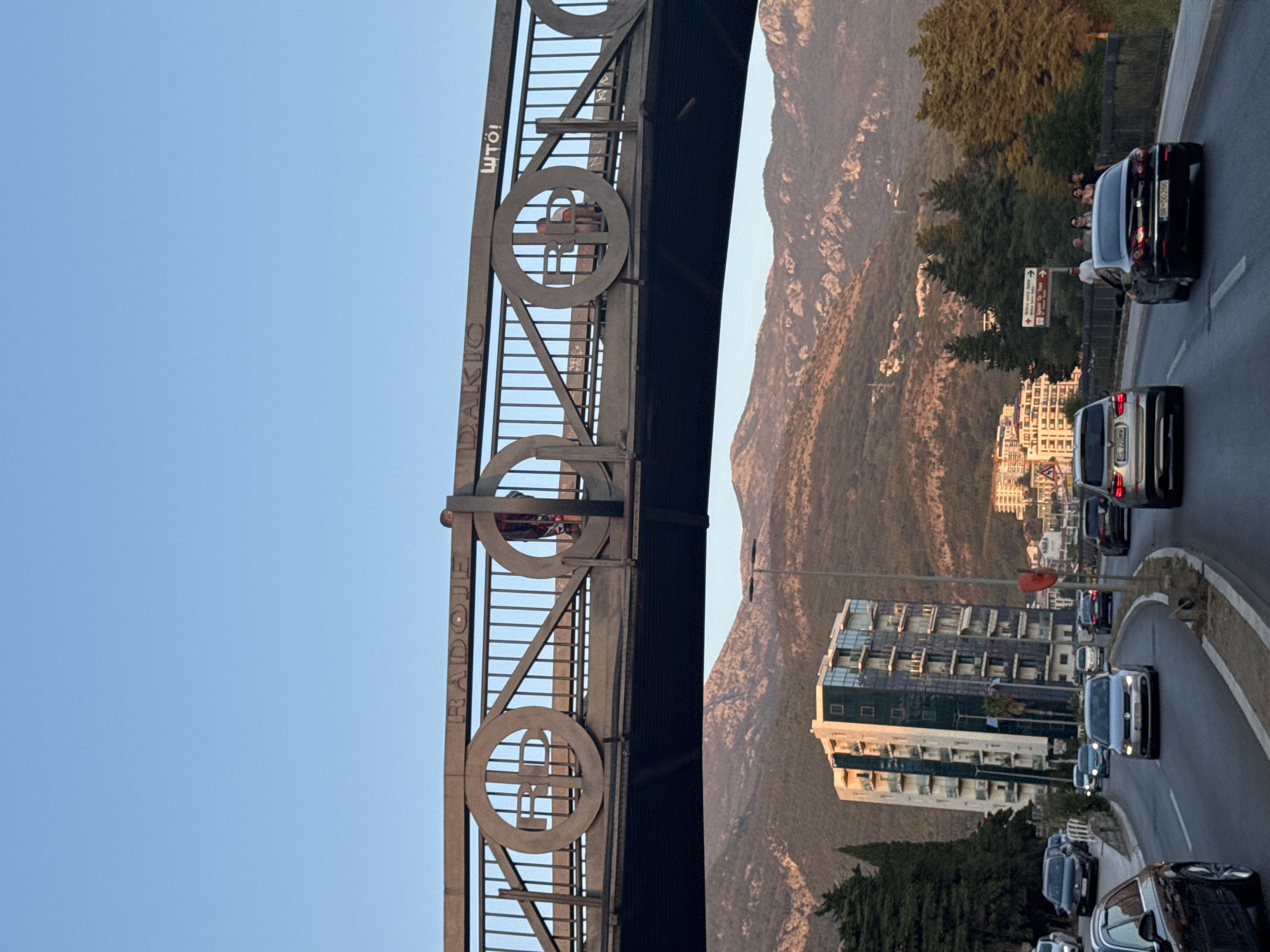 The image depicts a view from a car traveling on a road in Boreti, Montenegro, with a pedestrian bridge overhead. The bridge has the inscription "RADOJE DAKIC" and a symbol resembling a letter "R" inside a circle. Two individuals are visible on the bridge. The road is lined with trees and a modern, tall building with balconies. In the background, a mountainous landscape is illuminated by the golden light of what appears to be late afternoon or early evening. Multiple cars are visible on the road, some moving and some stationary. The sky is clear and blue.