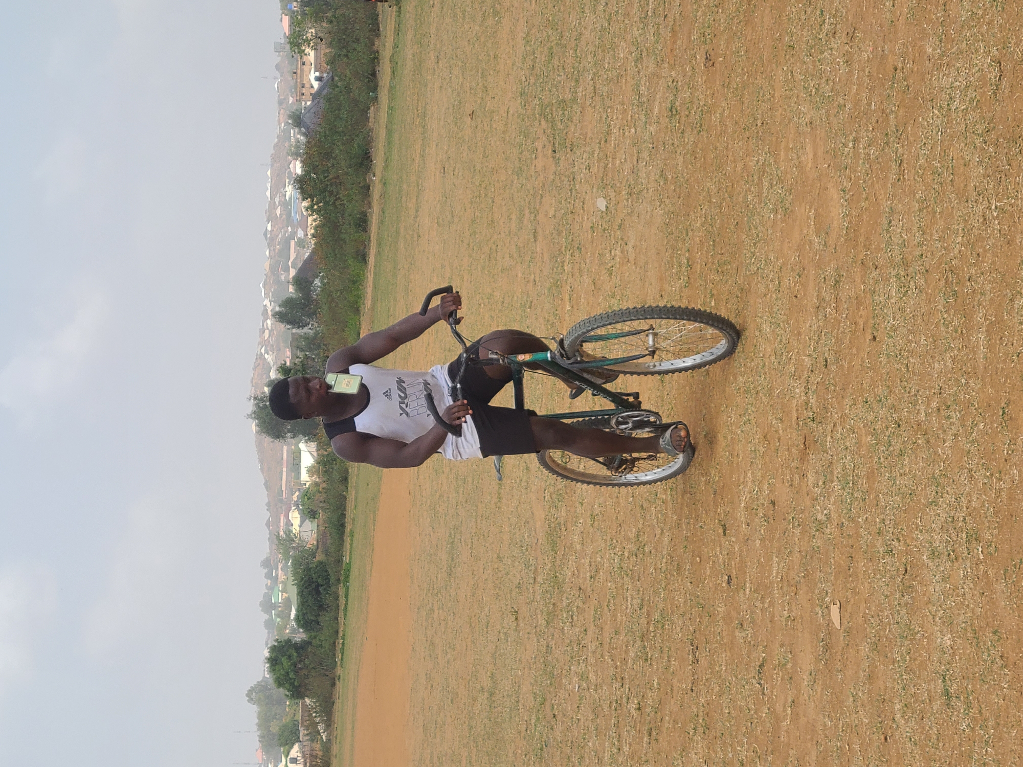 A man is sitting on a bicycle on a dry grass field in Barikin Jirga Summa, Nigeria. He is holding a phone to his ear. The town is visible in the background.
