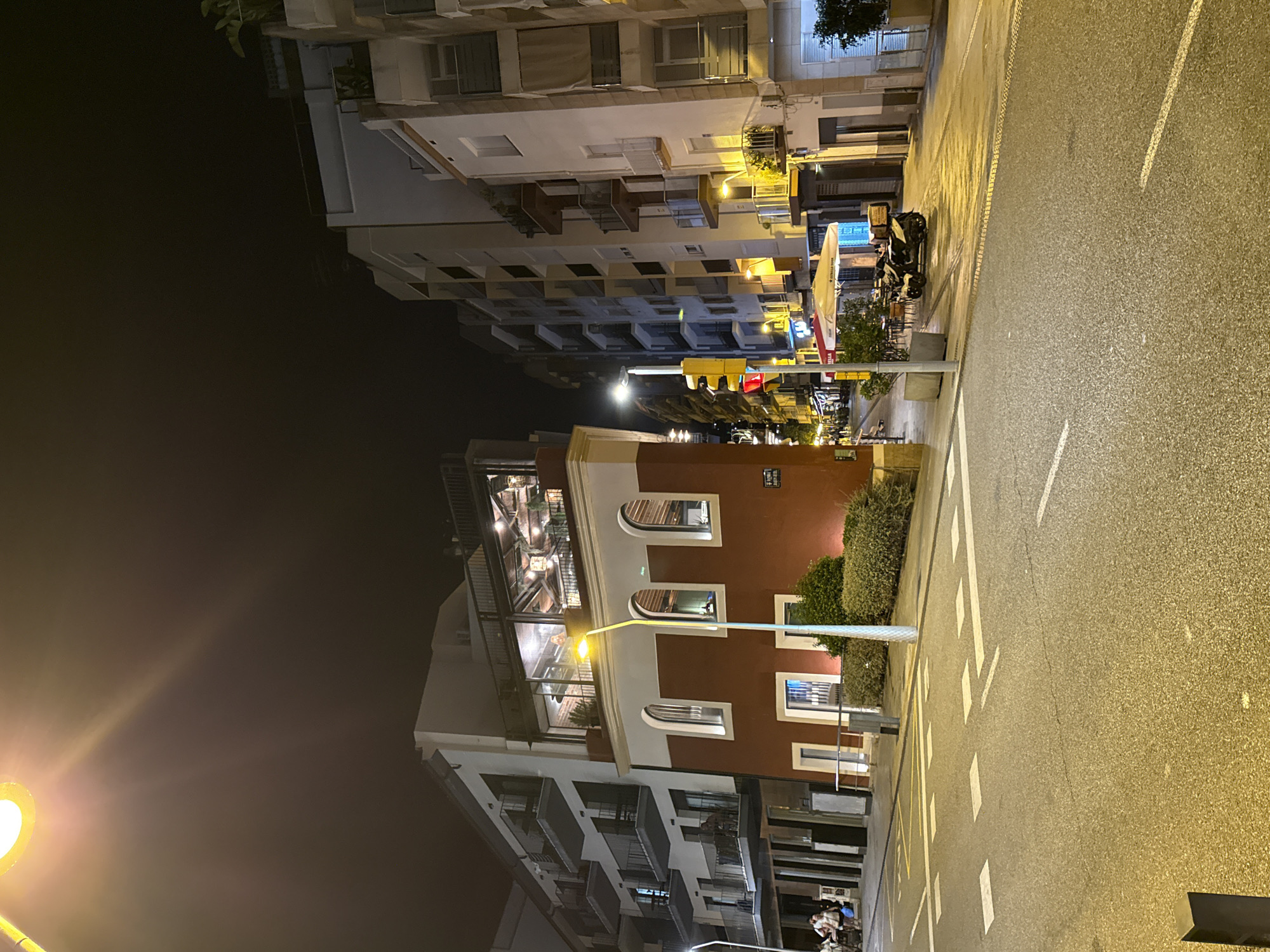 The image depicts a street scene at night in Blanes, Spain. On the right side of the street, there is a building with a distinctive red facade featuring arched windows on the upper floor and rectangular windows on the lower floor. Above the red section is a white building with balconies. A brightly lit restaurant or bar with large windows is visible on the upper floor, showcasing an interior with wooden beams and lighting.
A yellow street lamp illuminates the foreground and parts of the buildings. A traffic light is visible on the right side of the street, showing a red light. In the middle ground, there is a seating area with tables and umbrellas outside a building. A parked scooter with a delivery box is in front of the seating area. Further down the street, more buildings and streetlights are visible.
On the left side of the street, a multi-story building with balconies is present, with some people visible on the balconies. A few individuals are walking on the sidewalk in the distance. The street surface is asphalt with white road markings, including a pedestrian crossing. The overall environment suggests a lively urban area, possibly a commercial or entertainment district, during the evening hours.