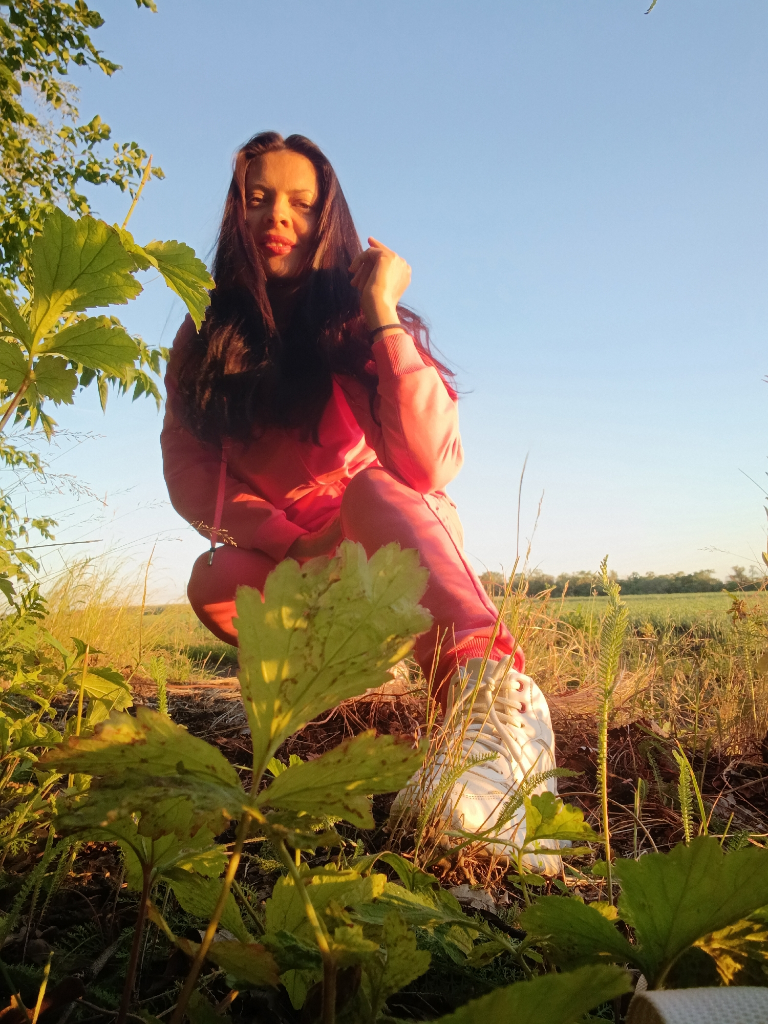 A young woman in a pink tracksuit and white sneakers is crouching in a field. She has long dark hair and is looking at the camera with red lipstick. She is holding one hand to her chin. In the foreground, there are large green leaves and tall grass. The background shows a green field and a clear blue sky. The lighting suggests it is either sunrise or sunset. The scene is likely outdoors, possibly in a rural area or park, given the natural surroundings. There is no indication of the city of Saratov, Russia, in the image.