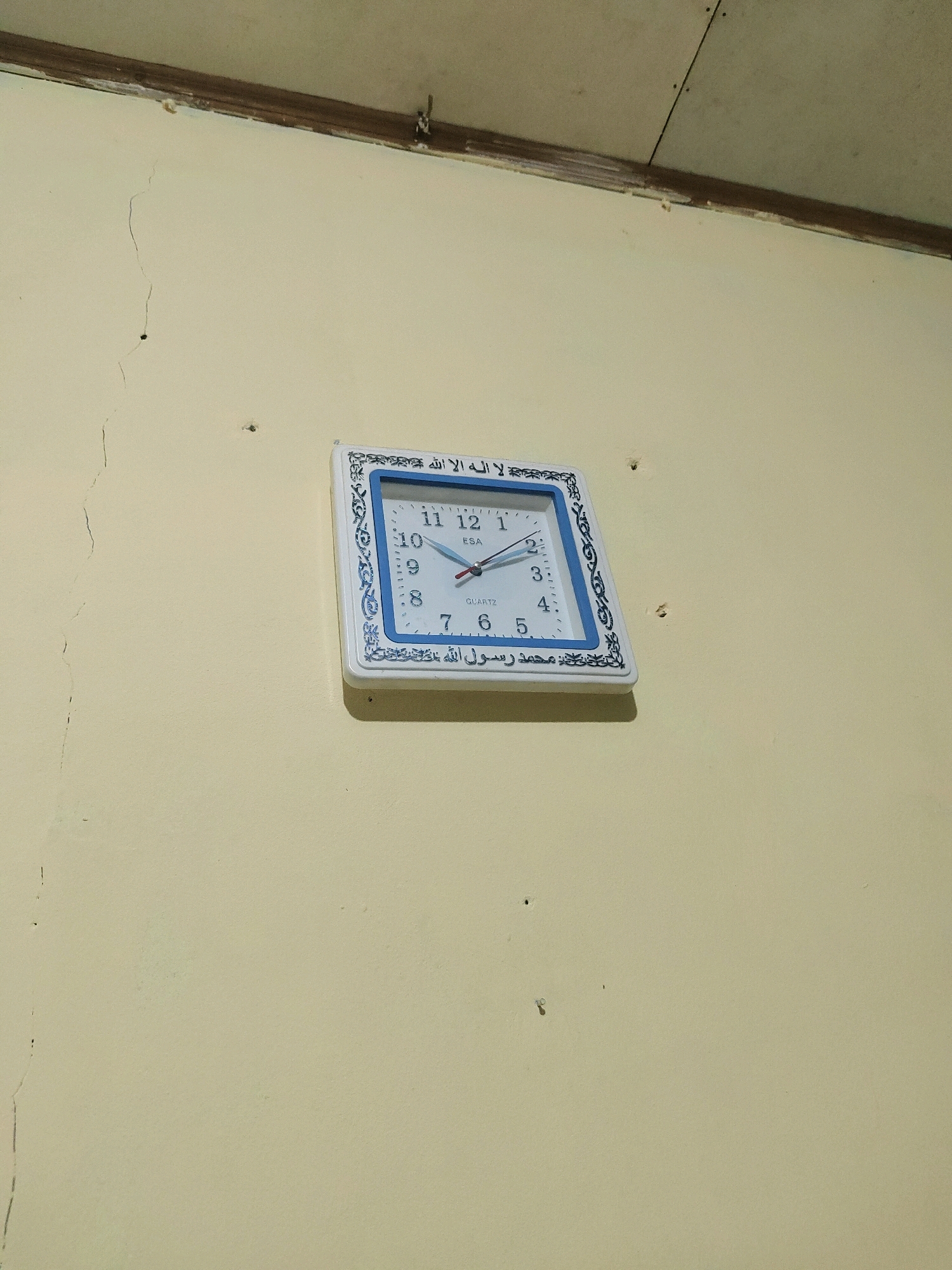 A square wall clock with a blue and white patterned frame hangs on a pale yellow wall. The clock face has black Arabic numerals and hands. The text on the clock is in Arabic script. The wall has several small cracks and holes. The ceiling is visible at the top of the image, showing exposed wooden beams and white panels. This scene is likely indoors, possibly in a residential or public building in Sukanagara, Indonesia.