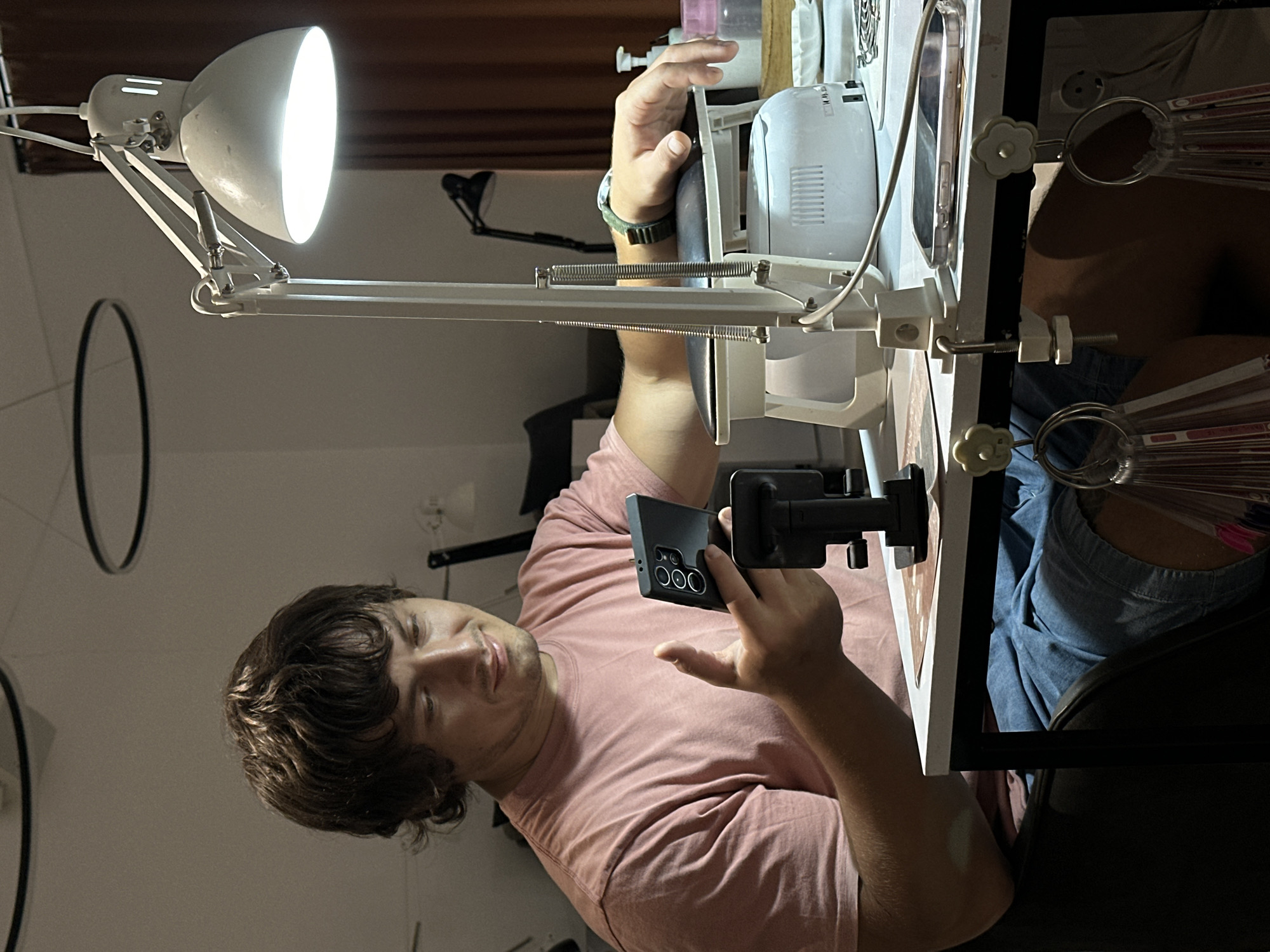 A man with brown, wavy hair is holding a black smartphone with three rear cameras. He is wearing a pink t-shirt and is positioned behind a white table with various items on it, including a white lamp, a white device, and what appears to be nail polish samples. The table is clamped to a dark chair or surface. The background is a dimly lit room. There is no definitive evidence to confirm the location is Kerobokan Kelod, Indonesia.