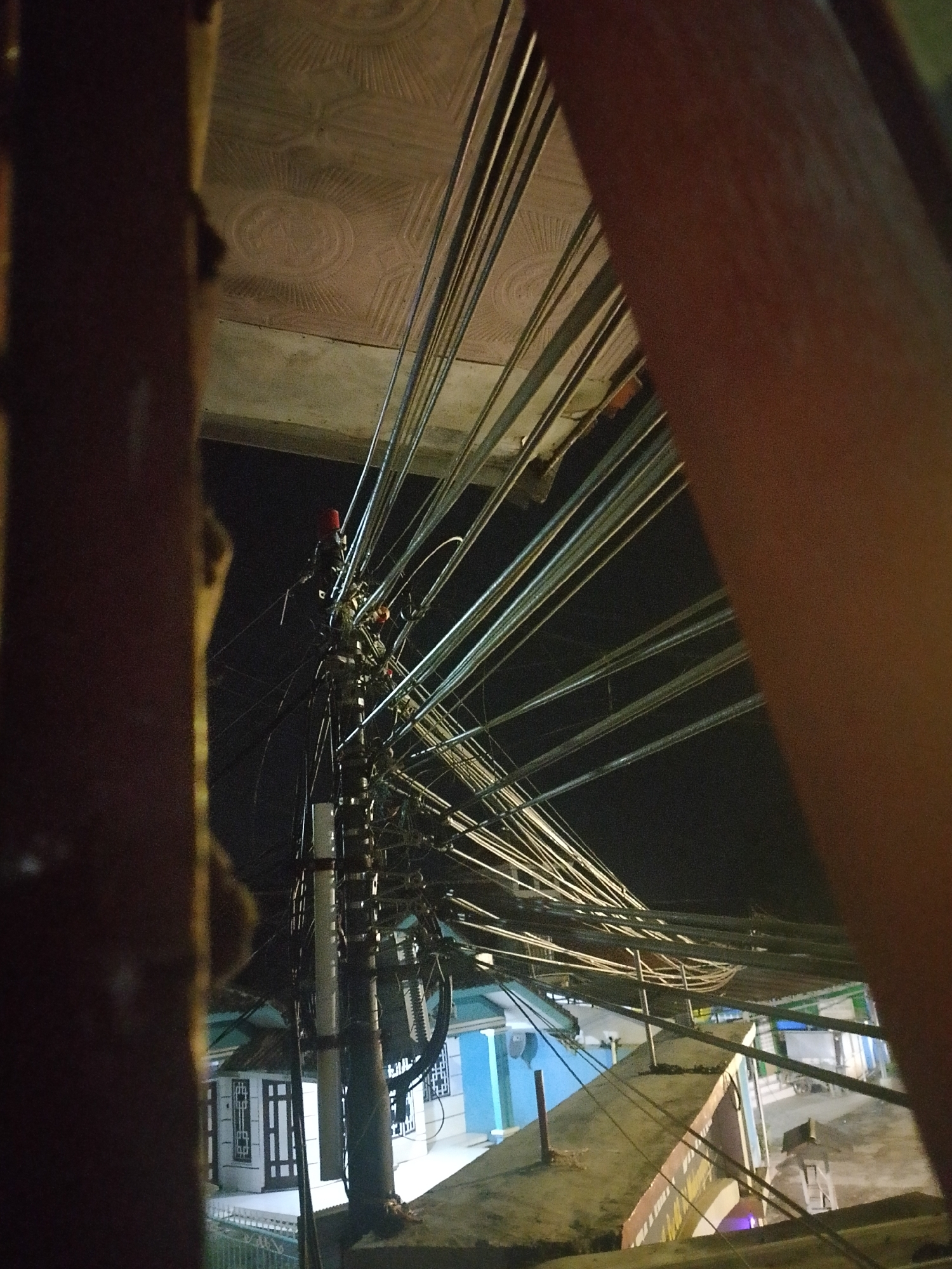 The image shows a dense network of overhead electrical and communication wires against a dark sky, illuminated by ambient light. In the foreground, a thick, vertical wooden beam on the left and a reddish-brown diagonal beam on the right partially obscure the view. The wires originate from a cluster near the top of a utility pole, which is visible in the mid-left section of the image. The pole is wrapped with what appear to be white insulation sleeves and has various attachments. Below the wires, several buildings with visible windows and doorways are present, some painted in light colors. The scene appears to be an urban or semi-urban environment, likely at night due to the dark sky and the artificial lighting. The general impression is one of a heavily wired area, typical of developing urban landscapes where infrastructure may be densely packed. The specific location in Mukapayung, Indonesia, suggests this scene is within a residential or commercial district of that area. There are no people visible in the image, and the focus is on the infrastructure and the surrounding buildings.