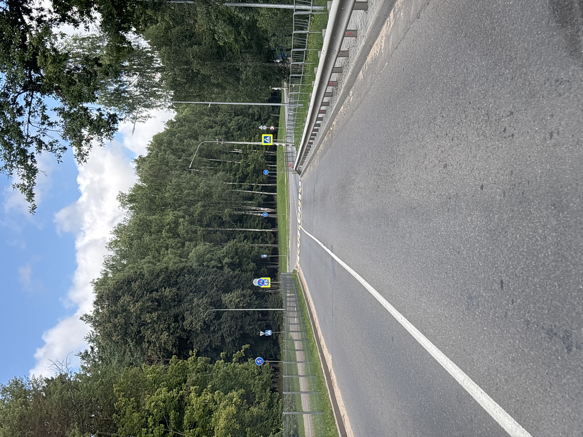This image shows a road in the Ждановіцкі сельскі Савет, Belarus. The road is paved and has a white line down the center. There are trees on either side of the road, and a fence along the right side. There are also several road signs, including a pedestrian crossing sign and a bicycle path sign. The sky is blue with some clouds. The scene appears to be a quiet, rural road.