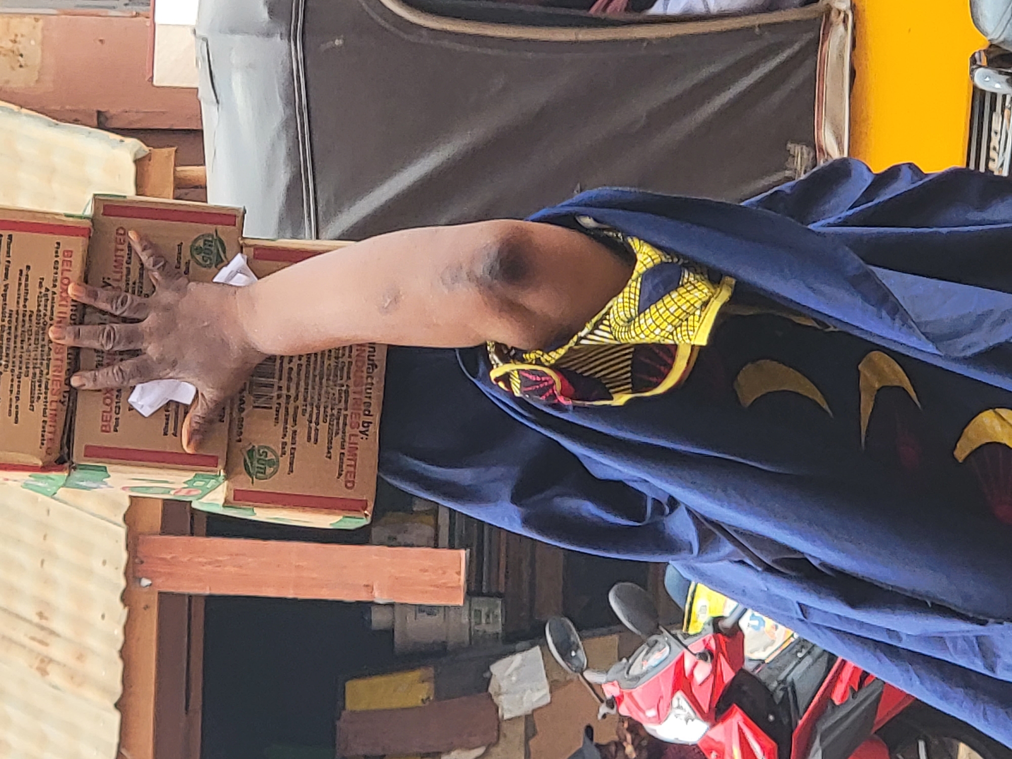 A woman's hand is resting on several Beloxxi Industries Limited boxes. The boxes are stacked on top of a wooden structure. The woman is wearing a blue garment with a yellow and red pattern. A red motorcycle is visible in the background. The scene appears to be outdoors, possibly in a market area in Maiburiji, Nigeria.