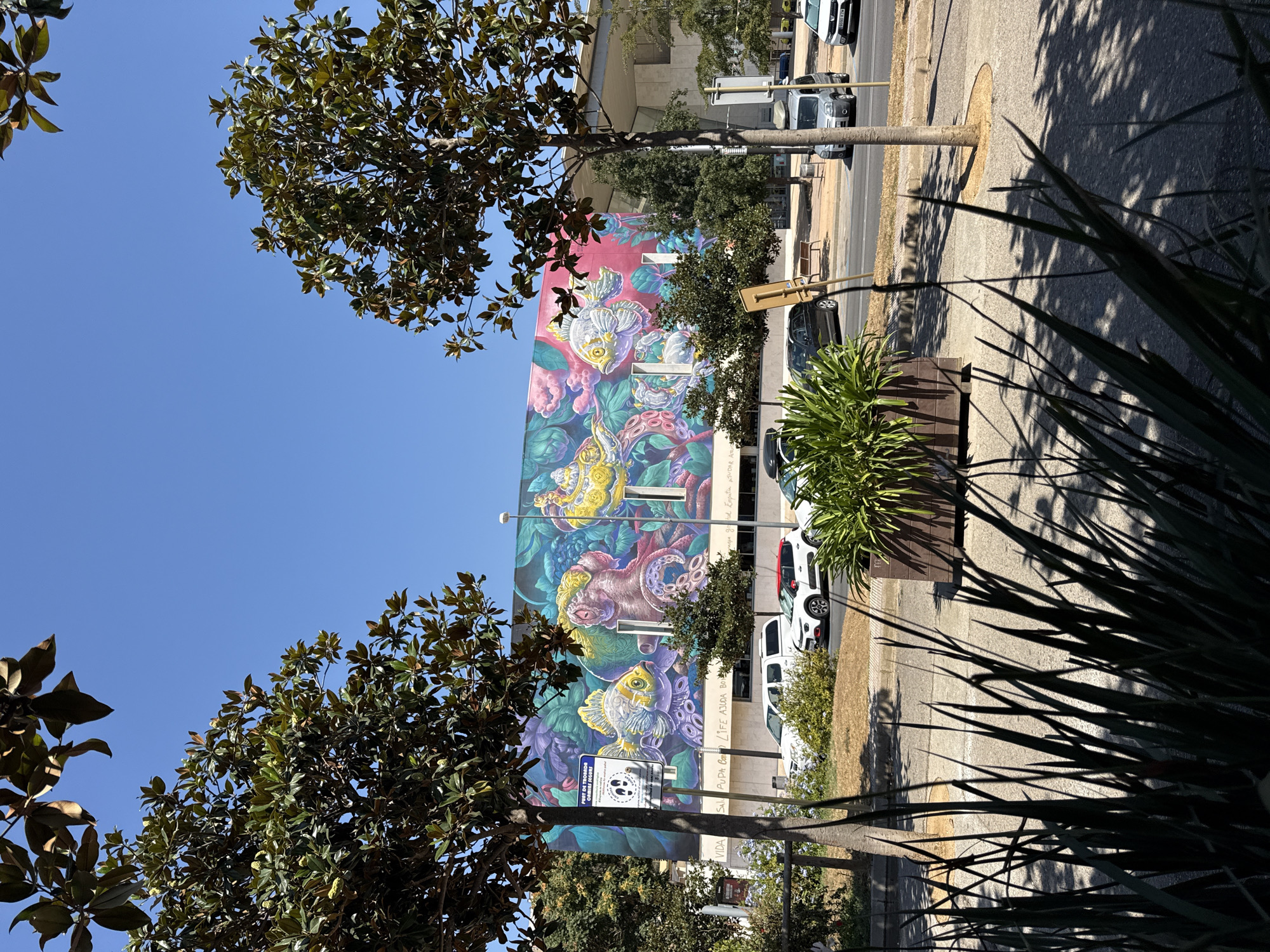 The image shows a mural on the side of a building in Blanes, Spain, depicting various sea creatures and plant life in vibrant colors. In the foreground, there are two trees with dark green leaves and a planter box filled with green foliage. A street runs in front of the building, with several cars parked and driving. The sky is clear and blue, indicating a sunny day. The mural features a large, yellow fish with intricate patterns, an octopus with pink and purple tentacles, and other colorful elements. A sign with walking figures and text is visible on the left side of the mural. The building itself appears to be modern with a concrete facade.